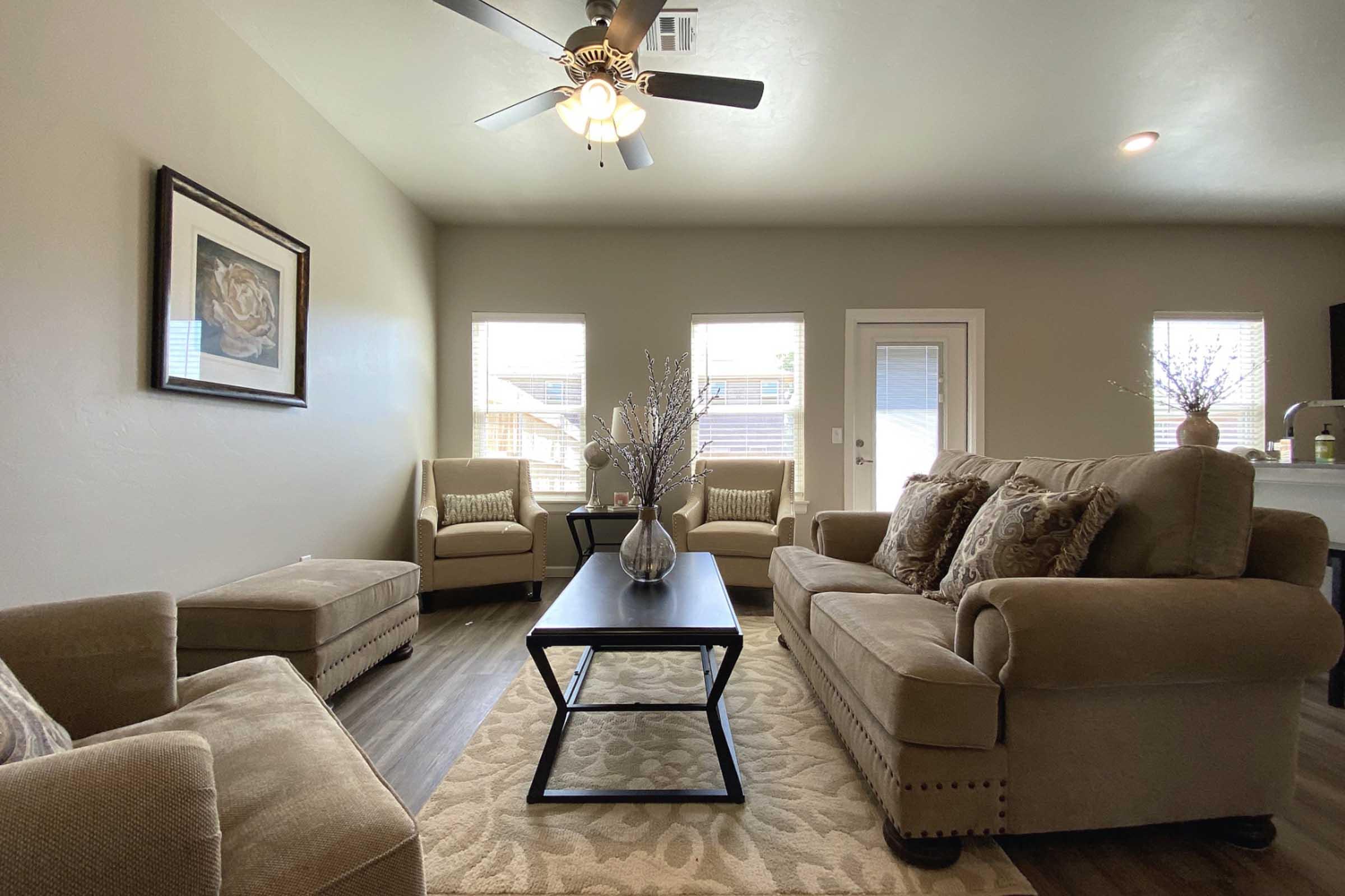 A cozy living room featuring a neutral color palette. There are two beige armchairs, a comfortable sofa with decorative cushions, and a wooden coffee table centered on a patterned rug. Sunlight streams through multiple windows, and a decorative vase with twigs is placed on the table, adding a touch of elegance.