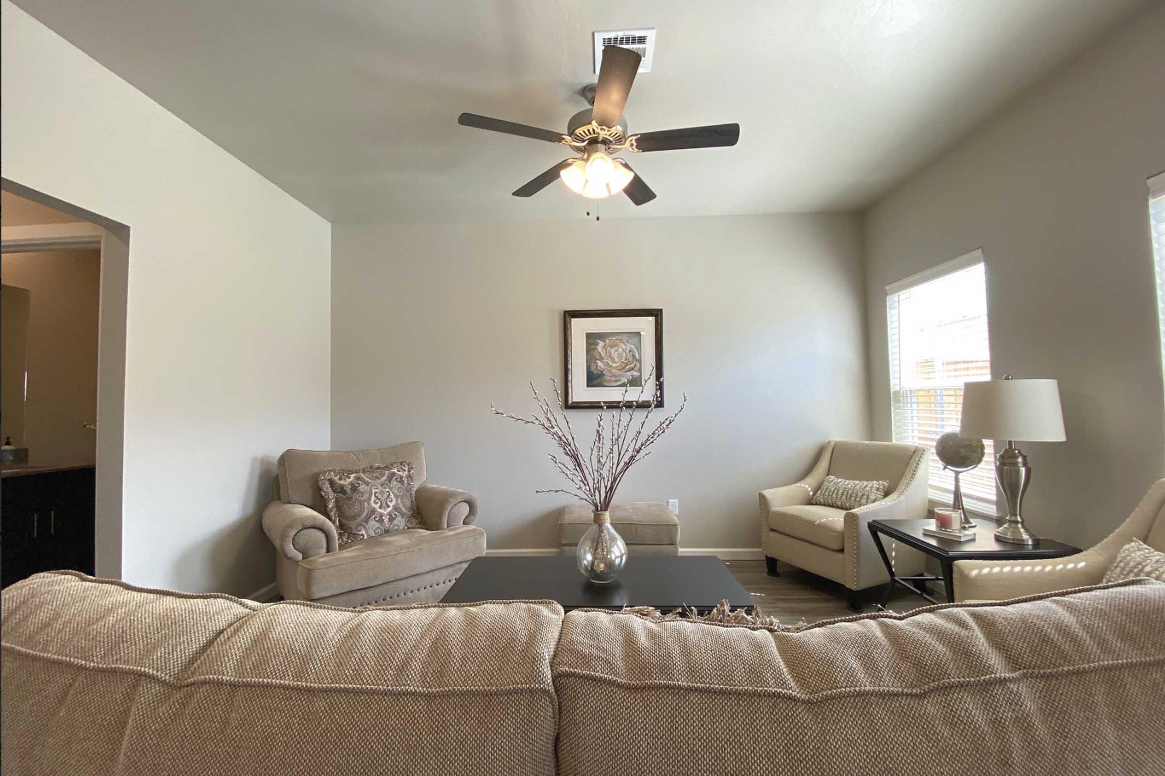 A cozy living room featuring a beige couch, an armchair, and a side chair. A coffee table sits in front, with a decorative vase and branches. A ceiling fan and two windows provide light, while a framed picture hangs on the wall, adding a touch of decor. The walls are light gray, creating a welcoming atmosphere.