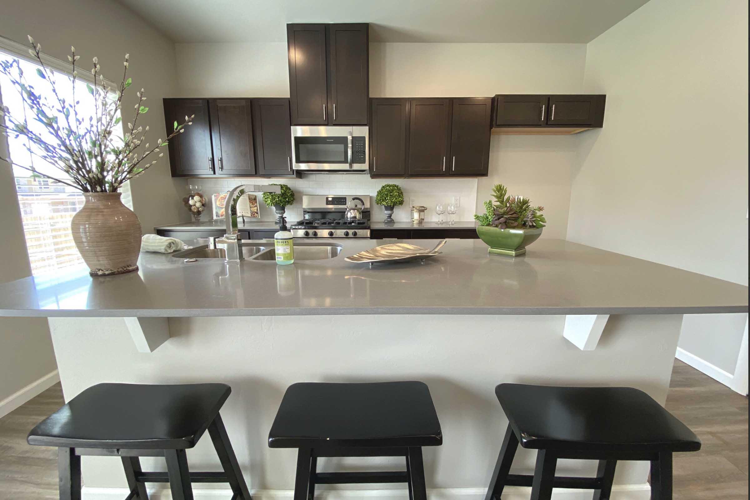 A modern kitchen featuring dark cabinetry, a stainless steel microwave, and a smooth countertop. In the foreground, three black bar stools are positioned at the counter. The countertop is decorated with a green plant and a vase of flowers, while a window allows natural light to brighten the space.