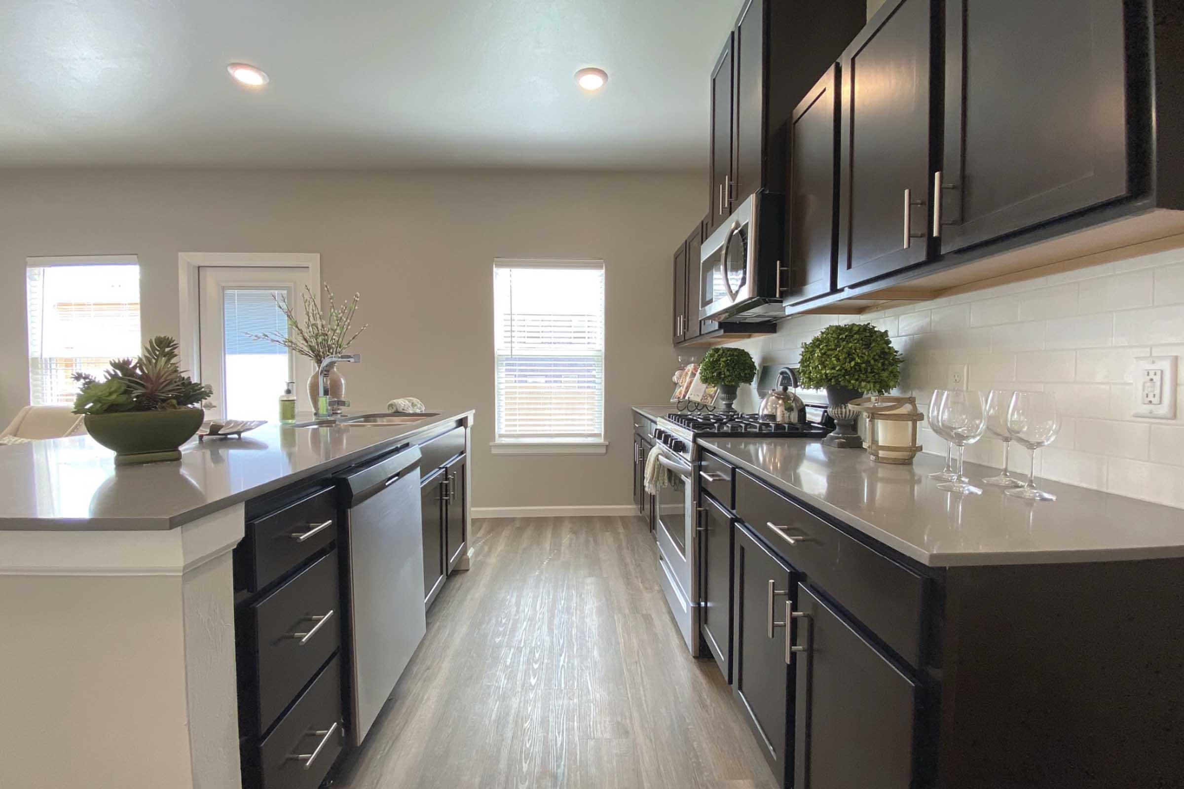 Modern kitchen interior featuring dark wood cabinetry, stainless steel appliances, and a light-colored countertops. A central island with a sink is visible, along with decorative plants. Natural light streams in through windows, creating a bright and inviting atmosphere.