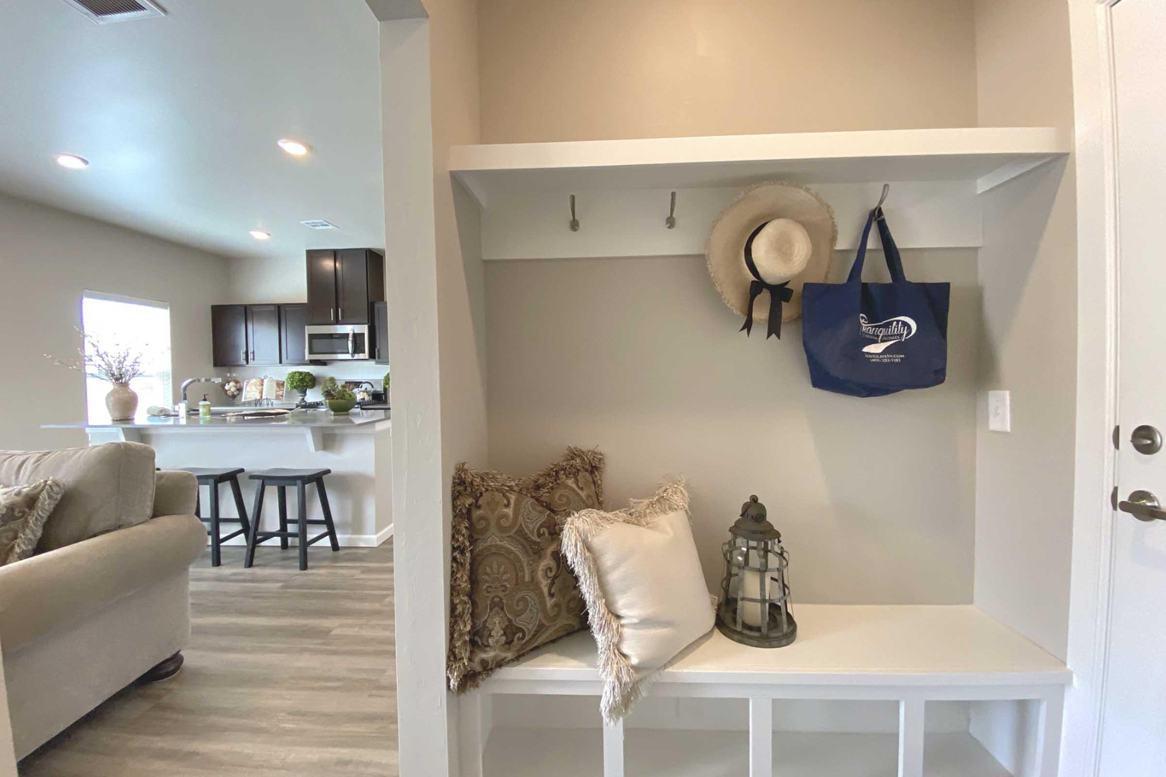A welcoming entryway featuring a white bench with decorative pillows, a lantern, and hooks for hanging items. A straw hat and a bag are hanging on the hooks, with a glimpse of a modern kitchen in the background, showcasing dark cabinets and a bar area with stools.