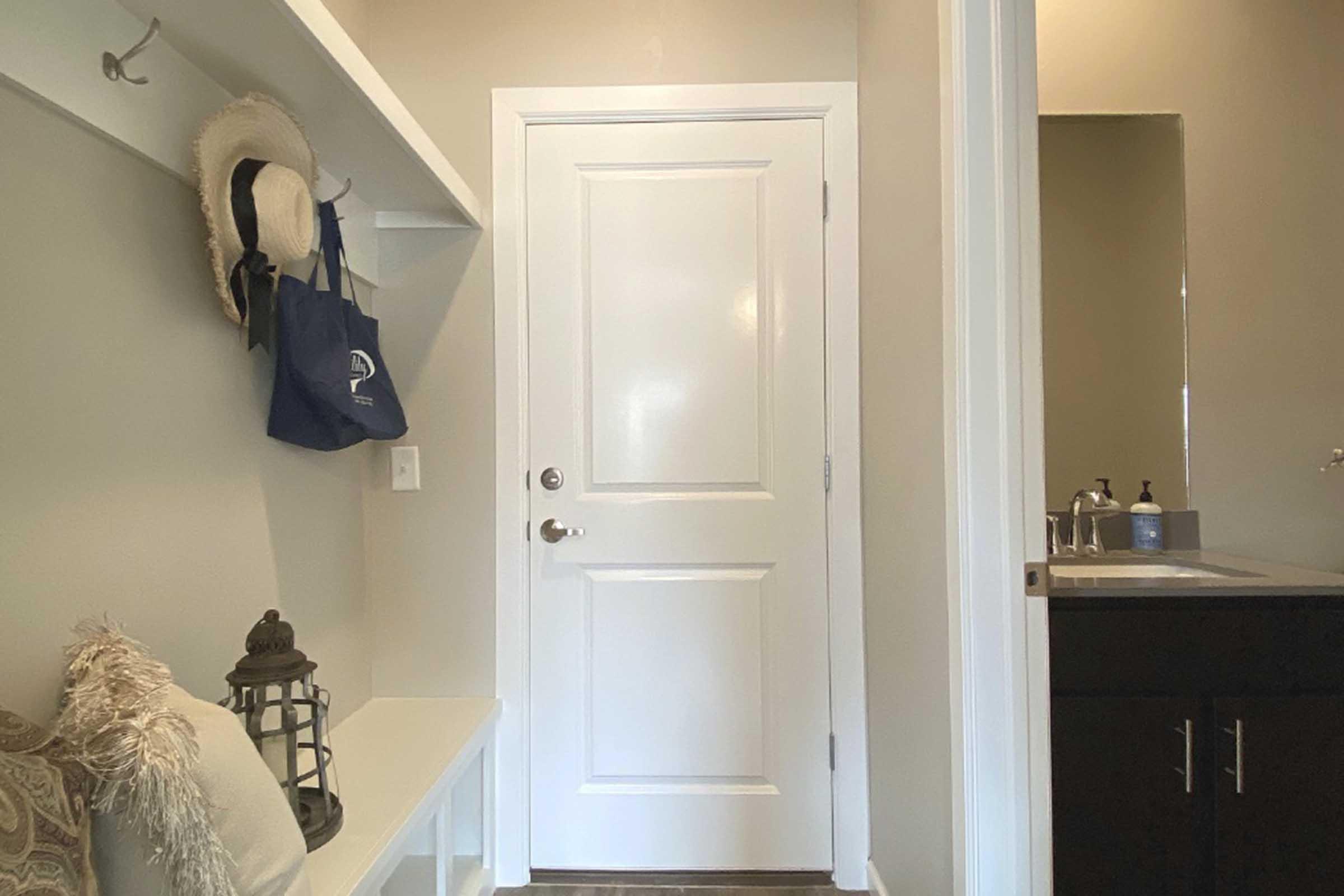 A hallway featuring a white door, a small entryway bench, and a wall shelf with a hat and tote bags. A decorative lantern is placed on the bench. The walls are painted in a soft beige, and there's a mirror visible in a small adjacent bathroom area.