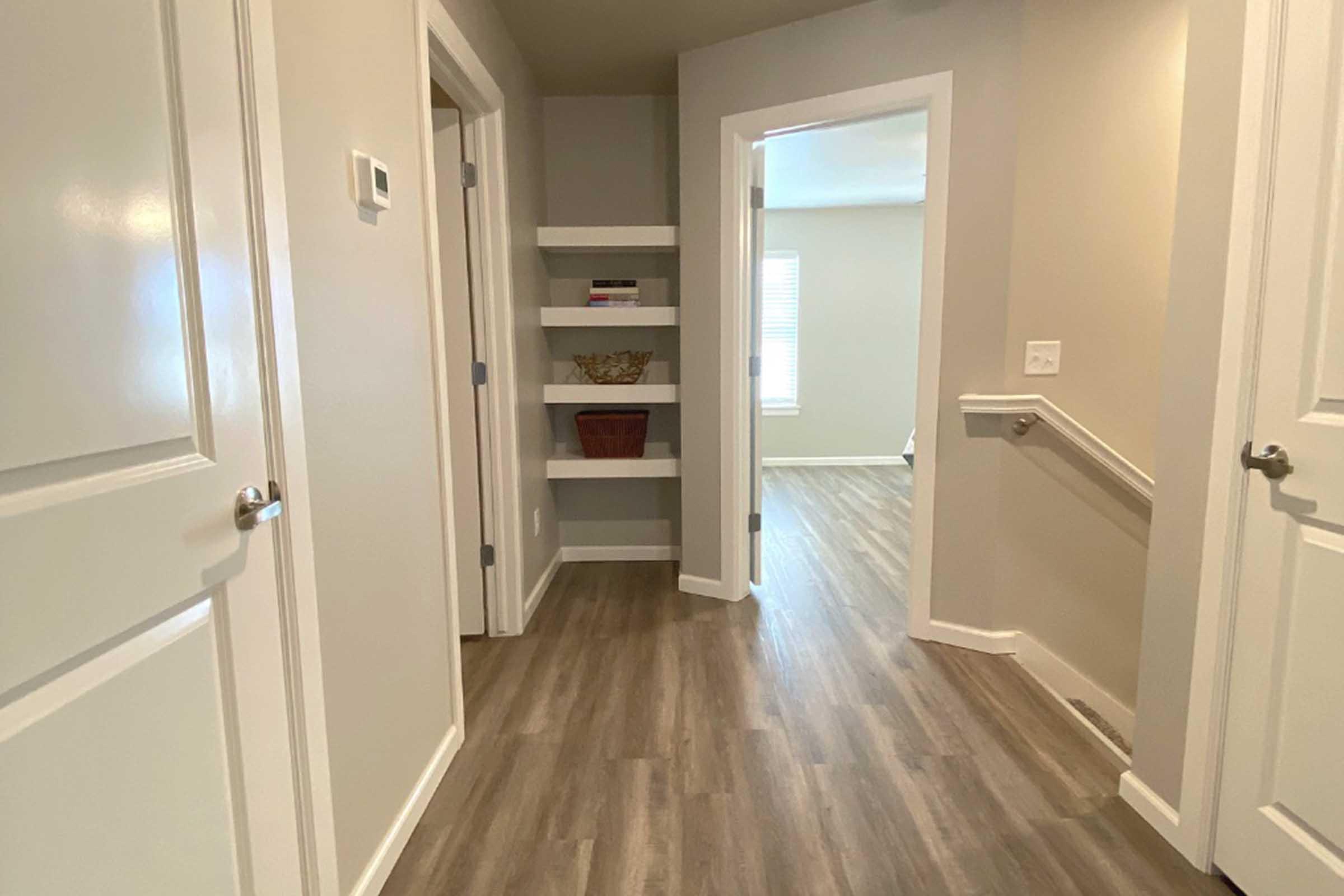 A well-lit hallway with light-colored walls and wooden flooring. On the left, there are two closed doors and a shelf with decorative items. A staircase is visible on the right side, leading to another room with a window at the far end, allowing natural light to enter.