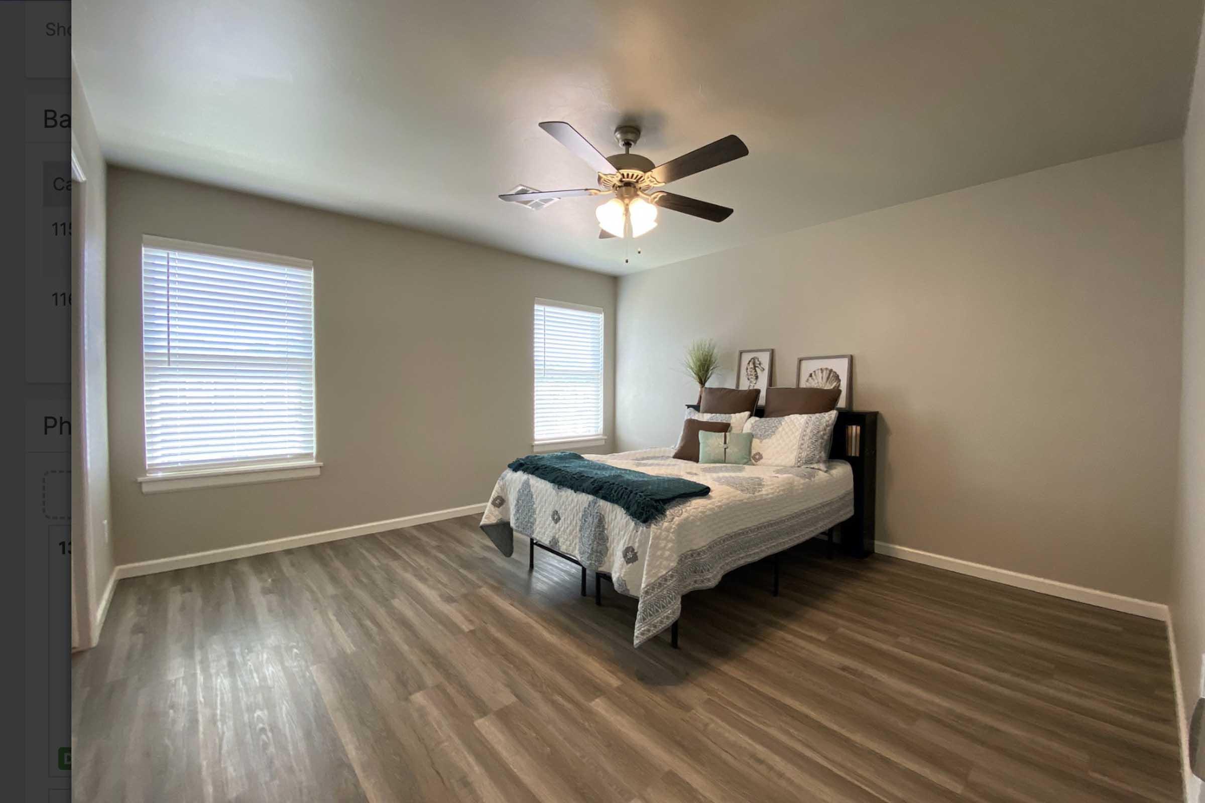 A well-lit bedroom featuring a bed with a teal and white patterned bedspread, two windows with blinds, and light wood flooring. There are framed pictures on the wall and a ceiling fan overhead, creating a cozy and inviting atmosphere.