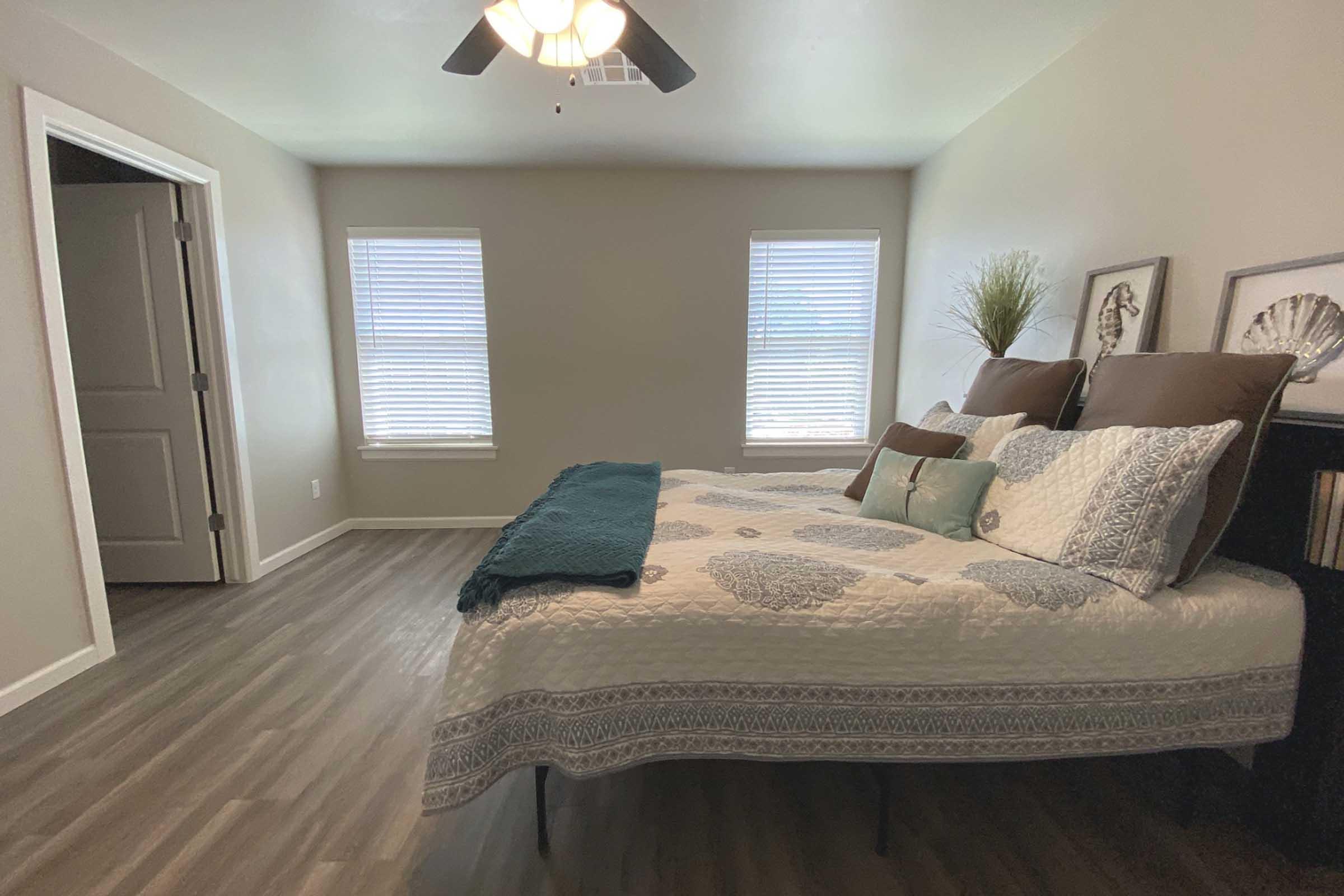 A cozy bedroom featuring a neatly made bed with a patterned quilt and accent pillows. Light-colored walls and wood flooring create a warm atmosphere. Two windows with blinds allow natural light to enter, and a door leads to another room. Decorative wall art and a plant add a touch of elegance.