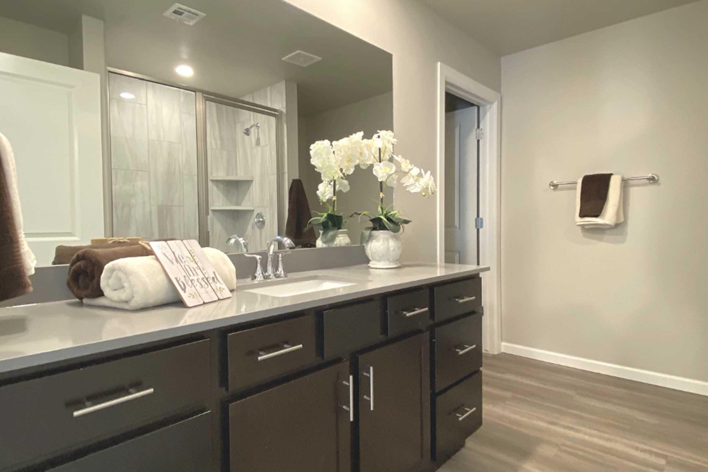 A modern bathroom featuring a double sink vanity with dark cabinets, a decorative vase of white orchids, neatly rolled towels, and a framed sign that says "Never stop dreaming." The wall has a towel rack, and the background includes a glass shower enclosure and light-colored walls.