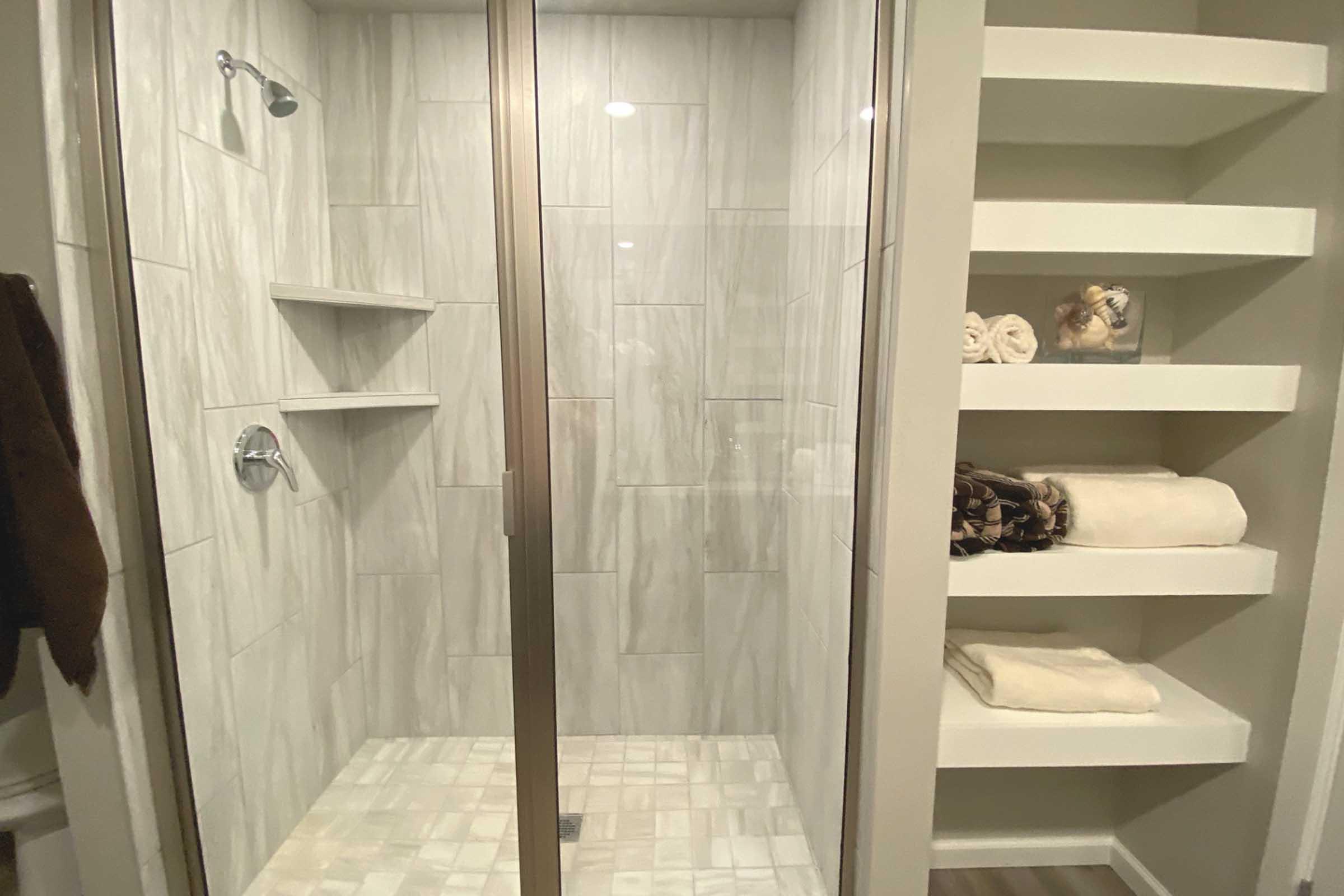 A modern, tiled shower with glass doors, featuring built-in shelves for toiletries. Adjacent to the shower is a storage area with neatly rolled towels and a decorative item on display. The overall design is sleek and contemporary, with neutral color tones.