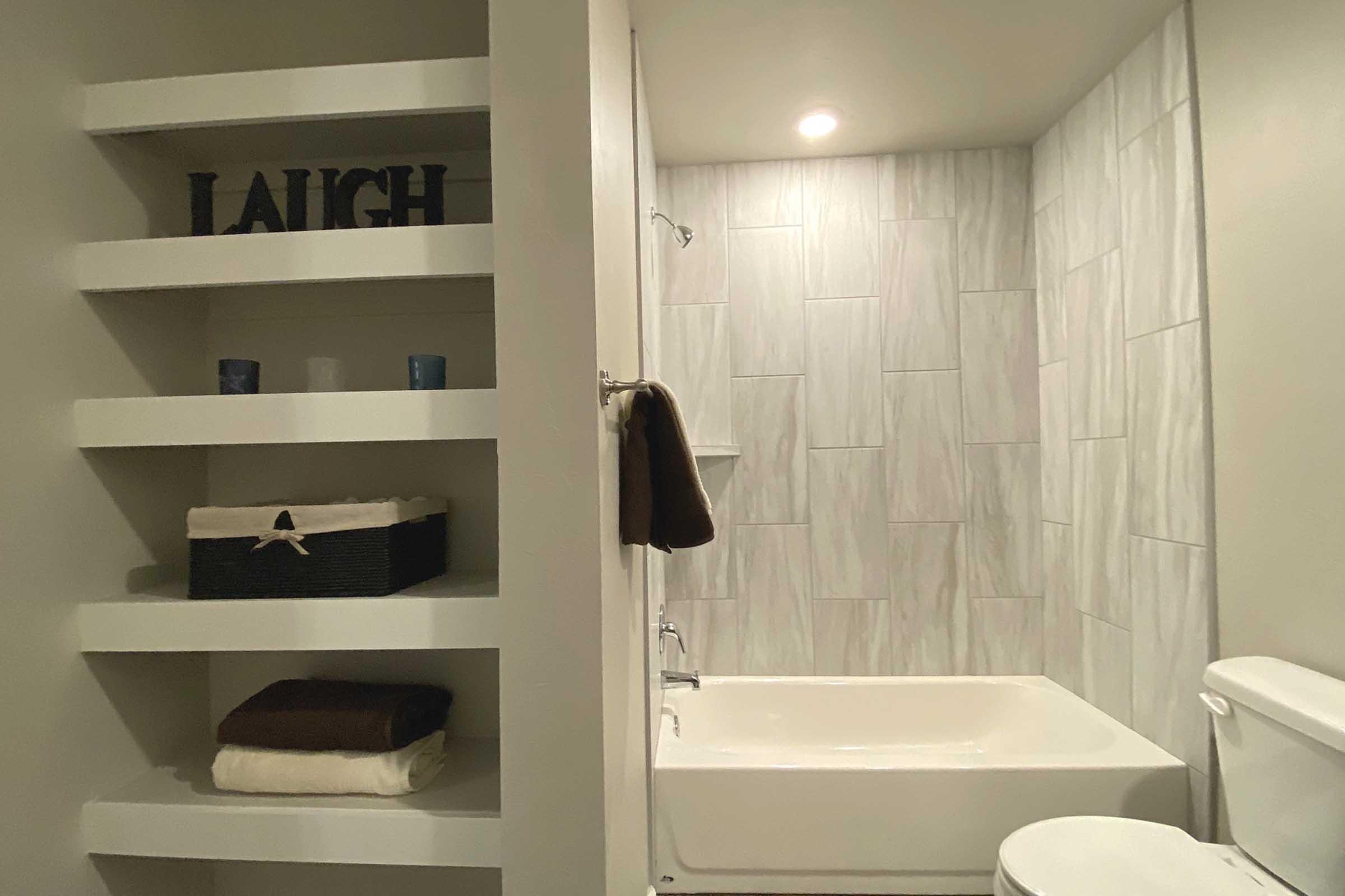 A clean and modern bathroom featuring a white tub, a toilet, and shelves. The shelves hold a "LAUGH" sign, decorative candles, and a woven basket. Soft towels are neatly stacked on the shelves, and the walls are adorned with light-colored, textured tiles. A warm light illuminates the space.