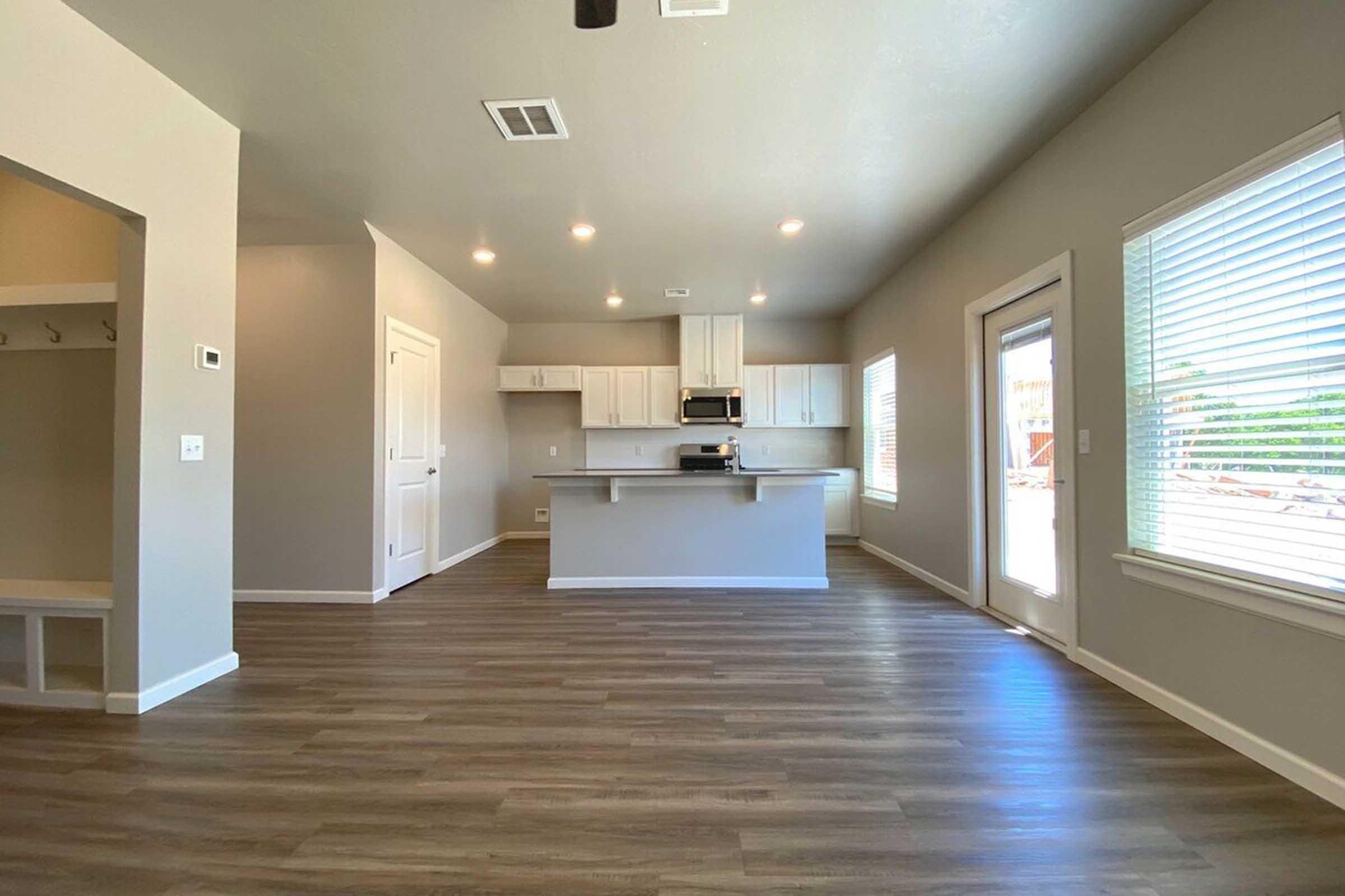 A bright, modern open-concept living space featuring a kitchen with white cabinets and stainless steel appliances. The room has large windows allowing natural light, light gray walls, and wood-look flooring. A door leads to an outdoor area, and a small entryway with coat hooks is visible.