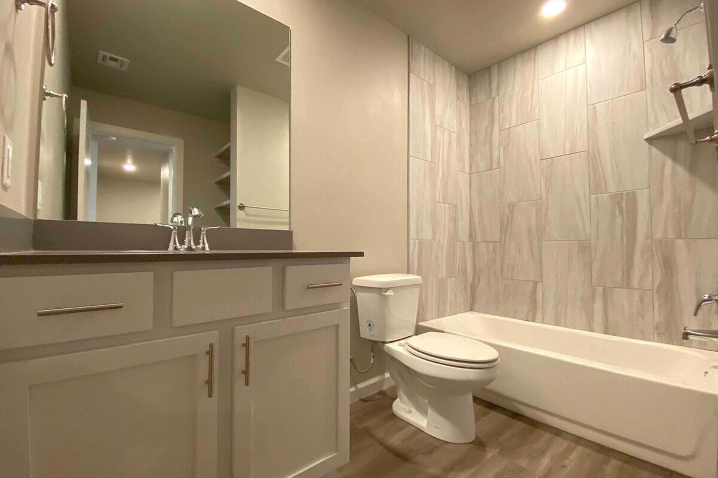 A modern bathroom featuring a white bathtub, a toilet, and a vanity with a gray countertop. The walls are lined with light-colored tiles, and there is a large mirror above the sink. Soft lighting illuminates the space, giving it a clean and contemporary look.