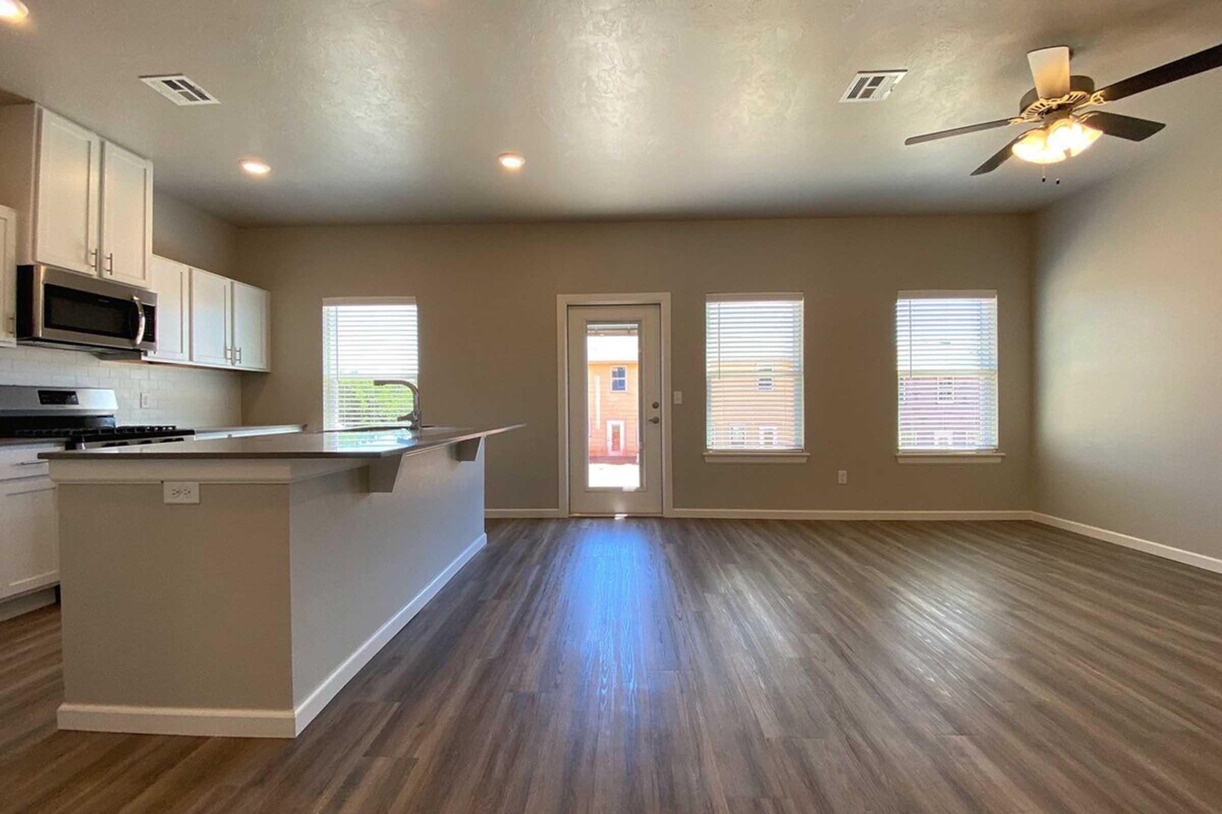 Interior view of a modern living space featuring a light-colored kitchen with stainless steel appliances, an island countertop, and an open layout. Large windows provide natural light, and the floor is finished with wood-like flooring. A patio door leads to an outdoor area.