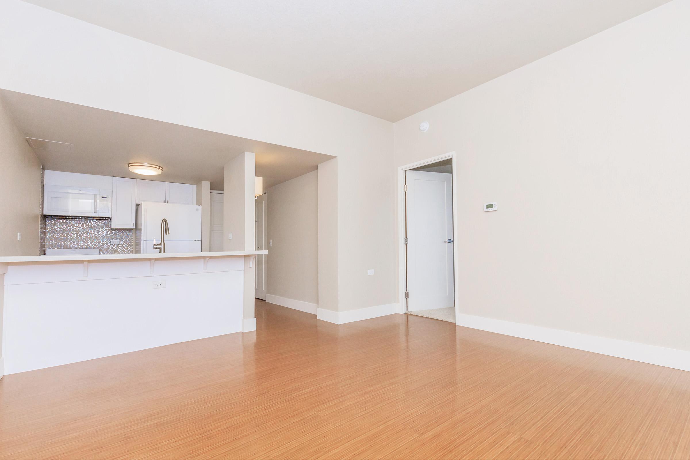 A modern, open-concept living space featuring light-colored walls, bamboo flooring, and a small kitchen area with white cabinetry in the background. A door leading to another room is visible, along with a thermostat mounted on the wall. Natural light fills the room, creating a bright atmosphere.