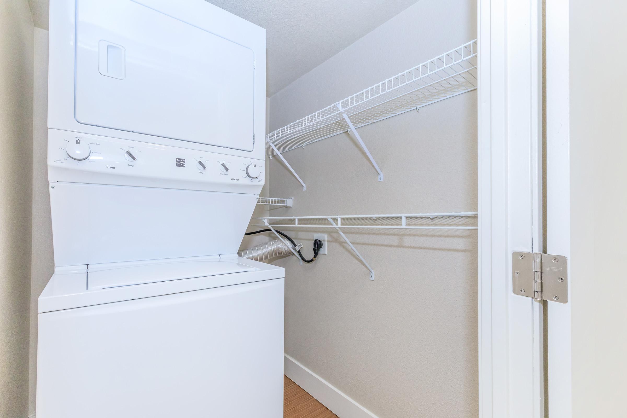 A compact laundry area featuring a stacked washer and dryer unit, with white wire shelving above for storage. The room has neutral-colored walls and a wooden floor, creating a clean and organized space for laundry tasks.