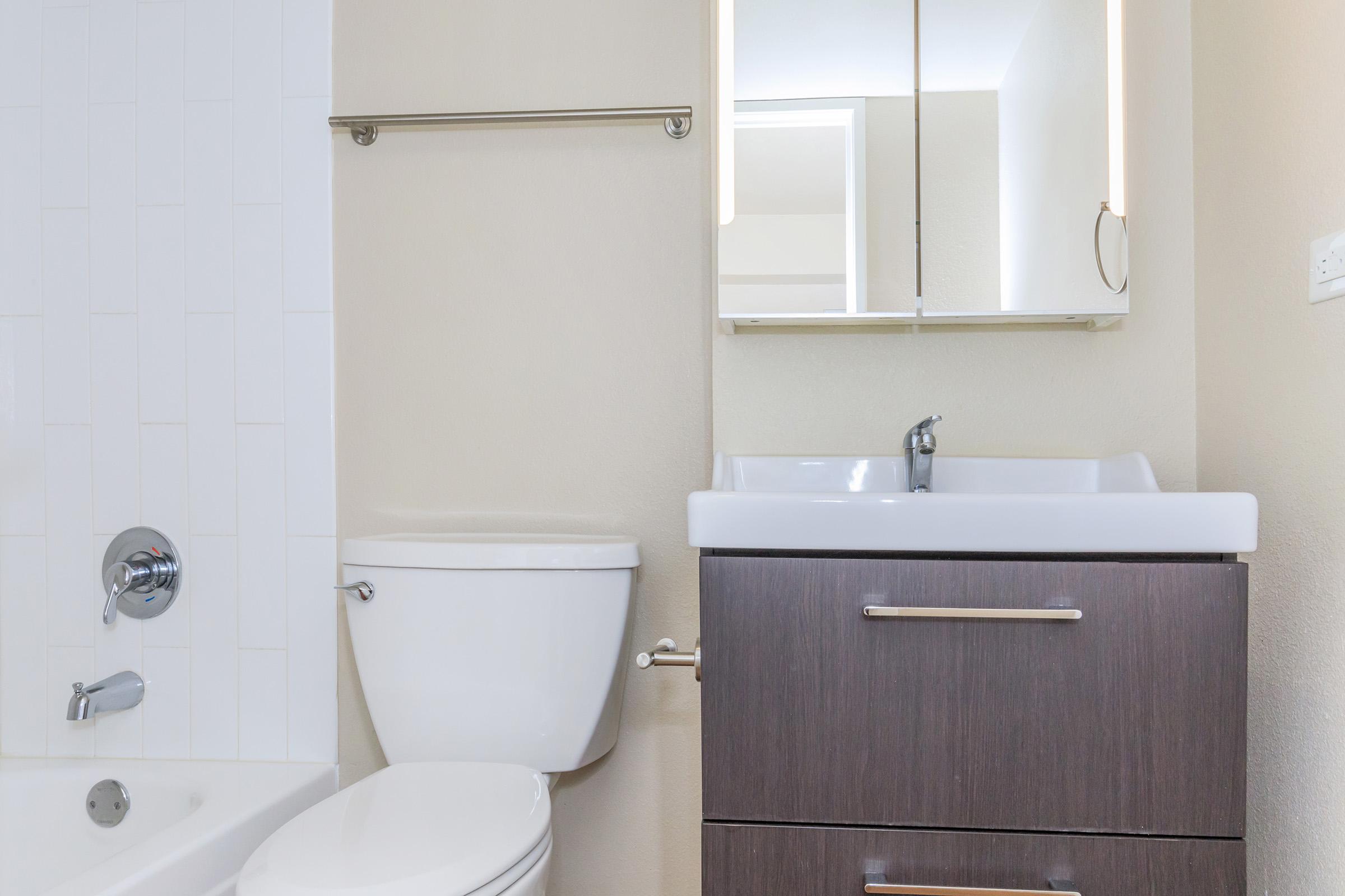 A modern bathroom featuring a white bathtub, wall-mounted faucet, and a sleek, dark wood vanity with a white sink. Above the sink, there is a mirror with integrated lighting. The walls are light-colored, creating a clean and bright atmosphere.