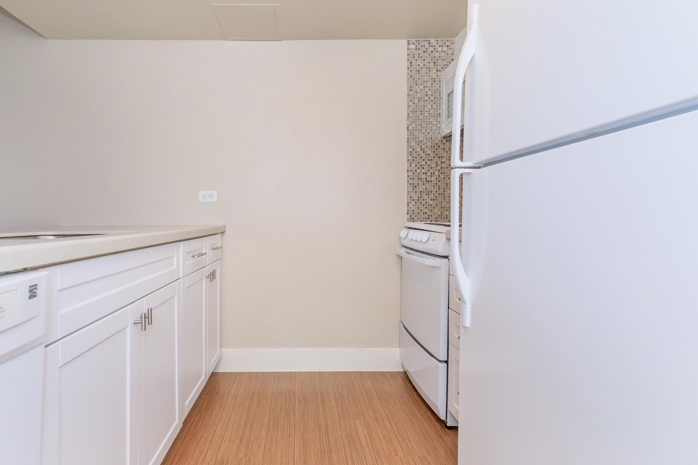 View of a modern kitchen featuring white cabinets, a countertop, a white stove, and a refrigerator. The walls have a light-colored finish, and a mosaic backsplash adds visual interest. The flooring is a light wood texture, creating a clean and airy atmosphere.