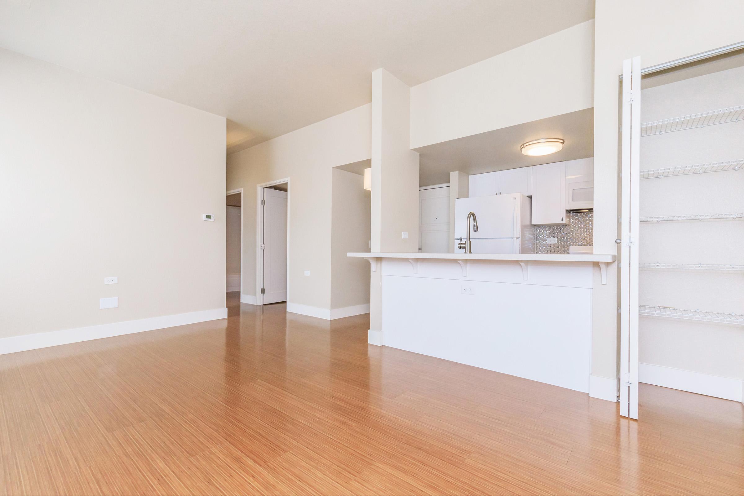 A spacious, modern living area with bamboo flooring, featuring a white kitchen island that separates the kitchen from the main space. Light-colored walls and ample natural light create an inviting atmosphere. There are doorways leading to other rooms in the background.
