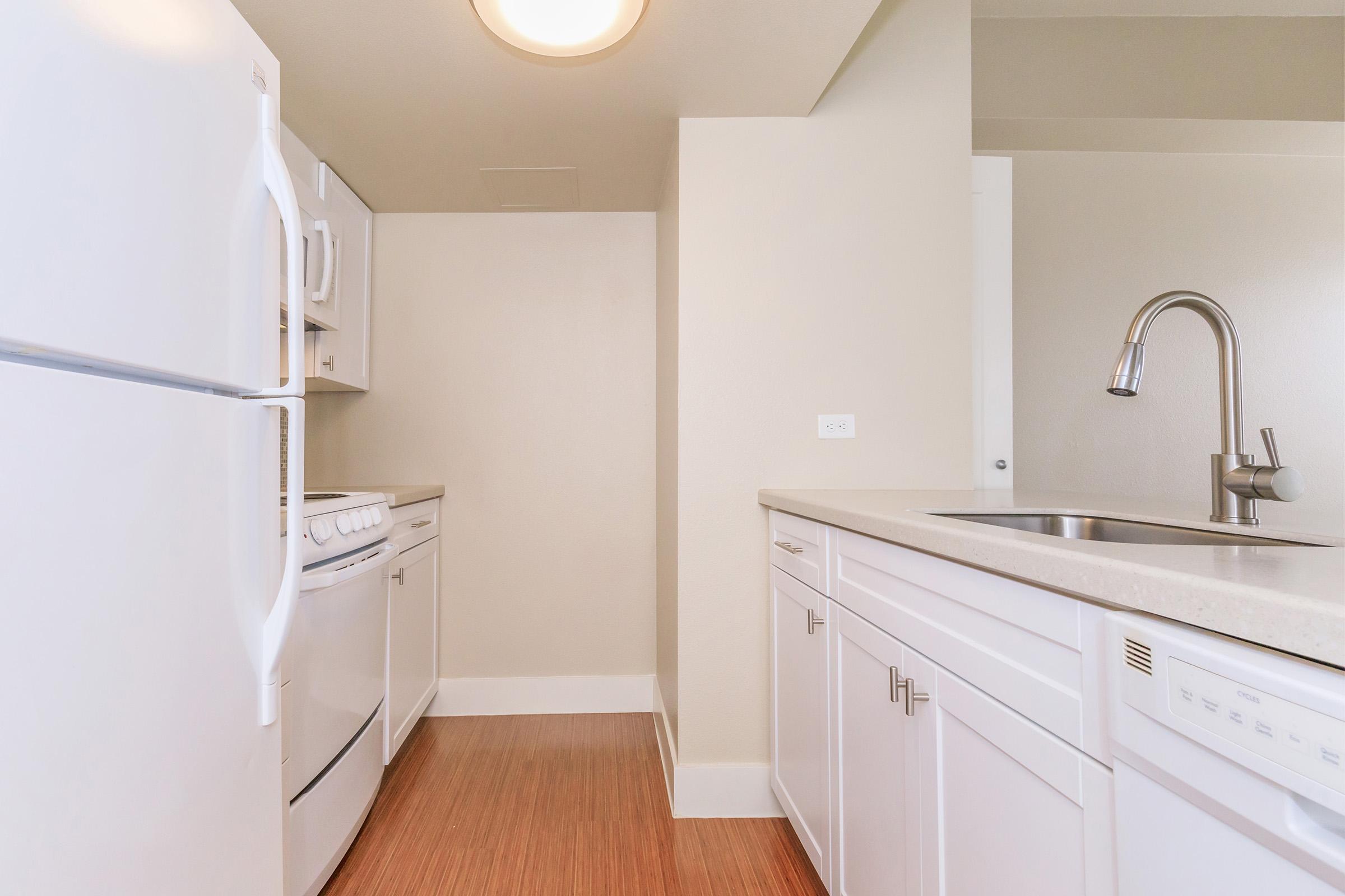 Modern kitchen featuring white cabinetry, a refrigerator, stove, and dishwasher. The space has a light-colored countertop and backsplash, with wood laminate flooring. Neutral walls and ample light create a bright and inviting atmosphere.
