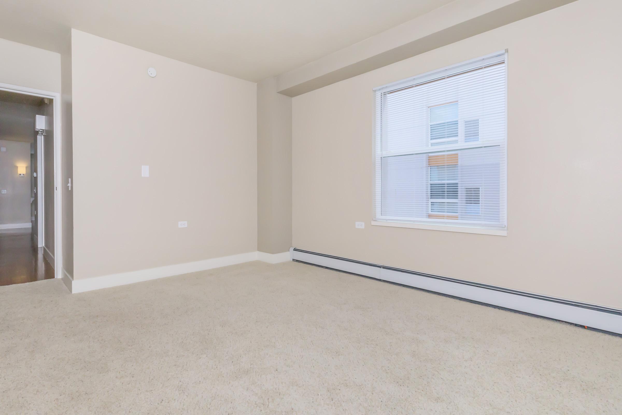 Empty room with beige walls and a light carpet. A single window with blinds on one side lets in natural light. The opposite wall has a door leading to a hallway. There are no furnishings or decorations in the space, creating a minimalistic and open atmosphere.