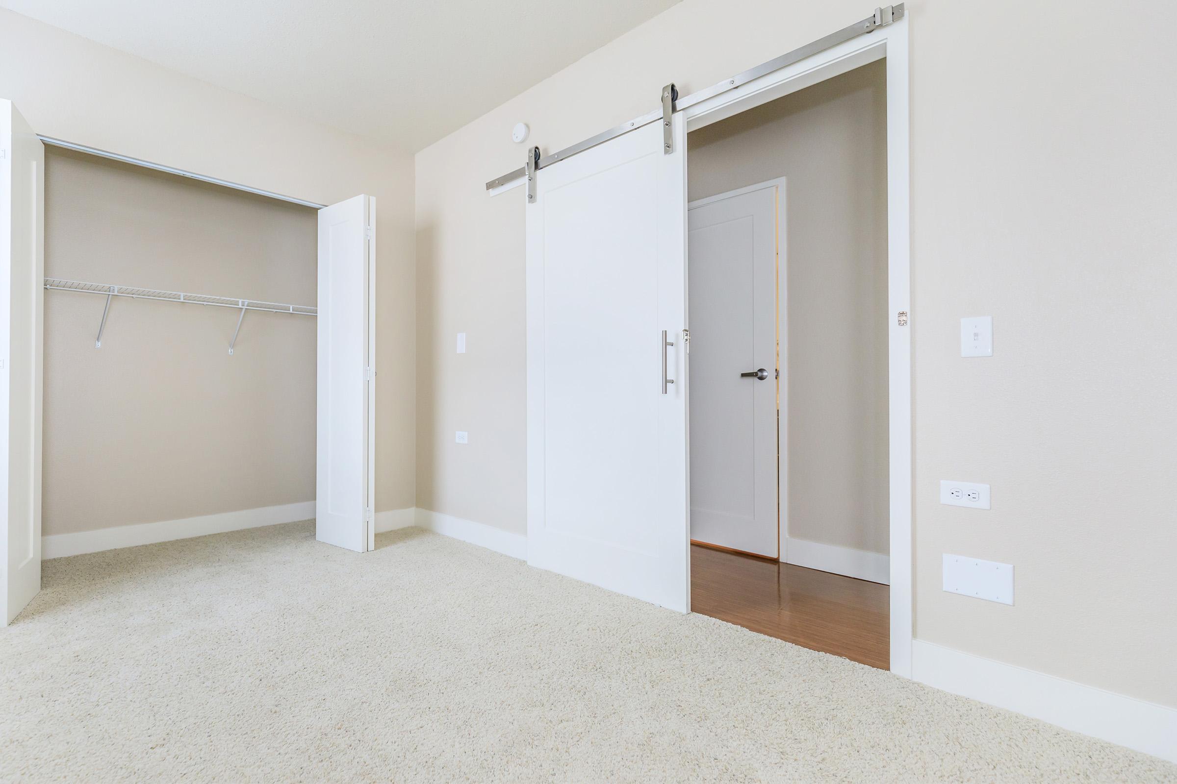 A well-lit, empty room featuring light-colored walls and carpet. There are two sliding closet doors on the left side and an open doorway on the right leading to another room. The overall aesthetic is modern and minimalistic, with a serene ambiance.