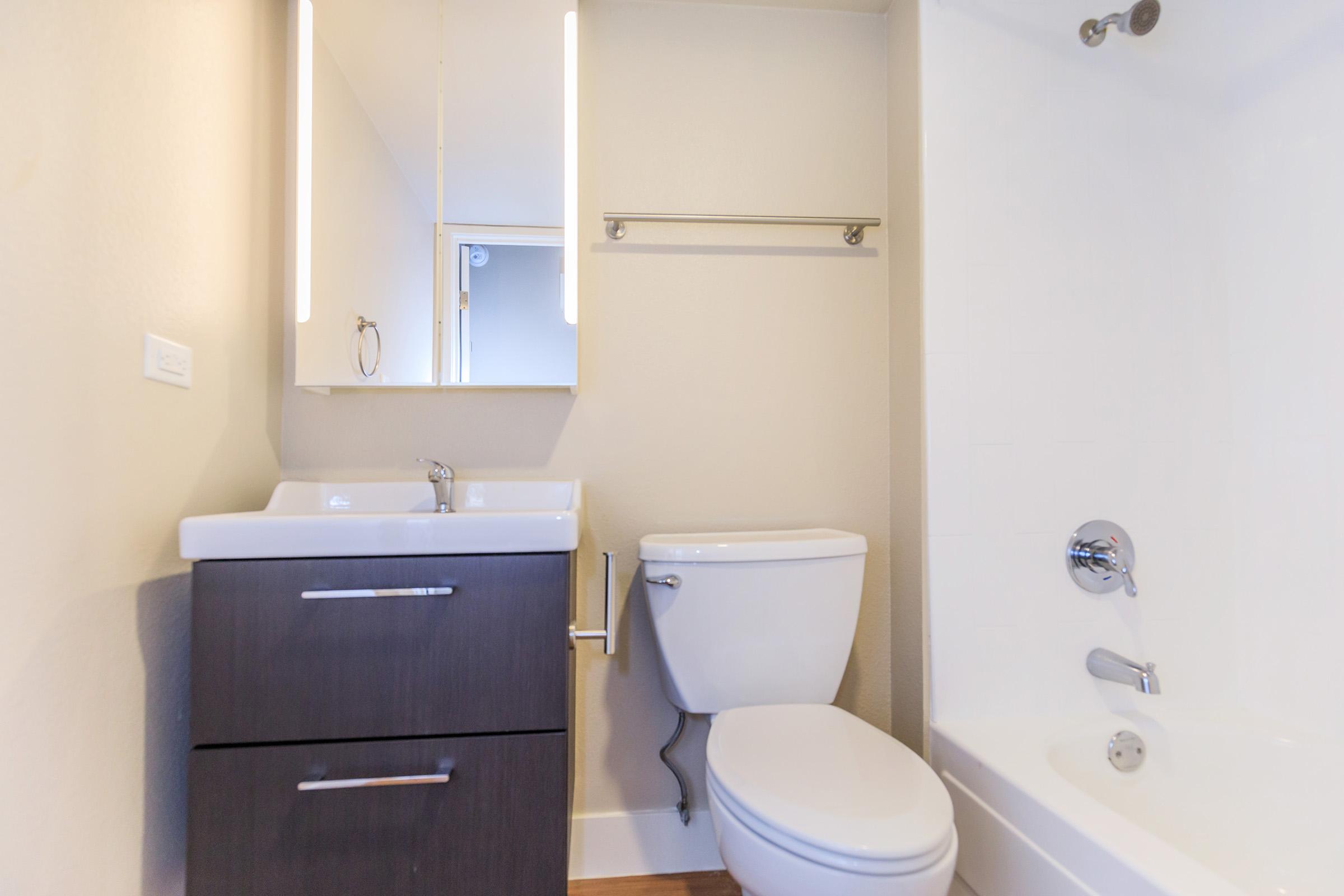 A small, modern bathroom featuring a white sink on a dark wood vanity, a toilet, and a bathtub with a shower. The walls are painted in a light color, and there is a mirror above the sink with built-in lighting. A towel bar is mounted above the toilet, enhancing the bathroom's functionality.