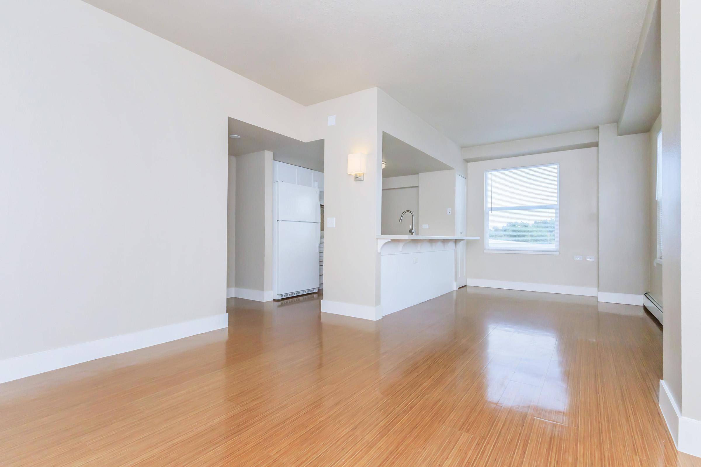 Spacious living area with bamboo flooring, featuring a kitchenette with a white refrigerator and a bar counter. Natural light streams in through a window. The walls are painted in neutral tones, creating a bright and airy atmosphere. Adjacent is an empty room for versatile use.