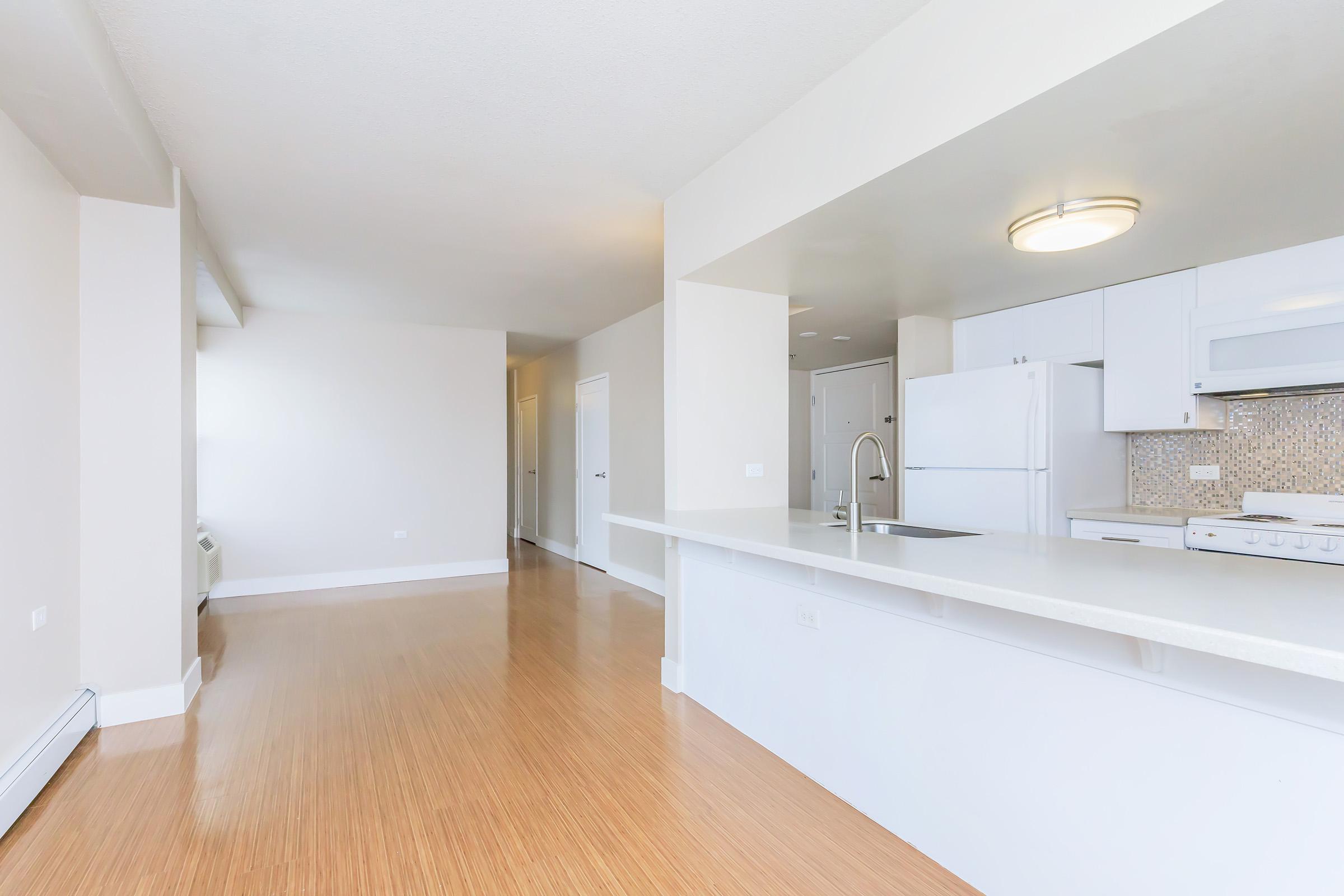 Bright and spacious open-concept apartment interior featuring a modern kitchen with white cabinetry, a light countertop, and a stylish backsplash. The living area has bamboo flooring and large windows, providing plenty of natural light. Simple, neutral decor enhances the inviting atmosphere.