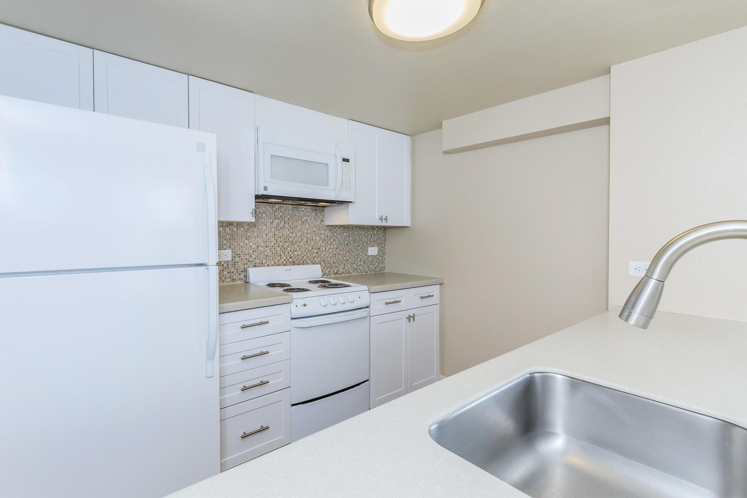 Modern kitchen featuring white cabinetry, a large refrigerator, and a microwave above the stove. The countertop is light-colored with a stainless steel sink. A neutral wall complements the space, and mosaic tiles add a decorative touch in the background. Bright lighting enhances the overall appearance.
