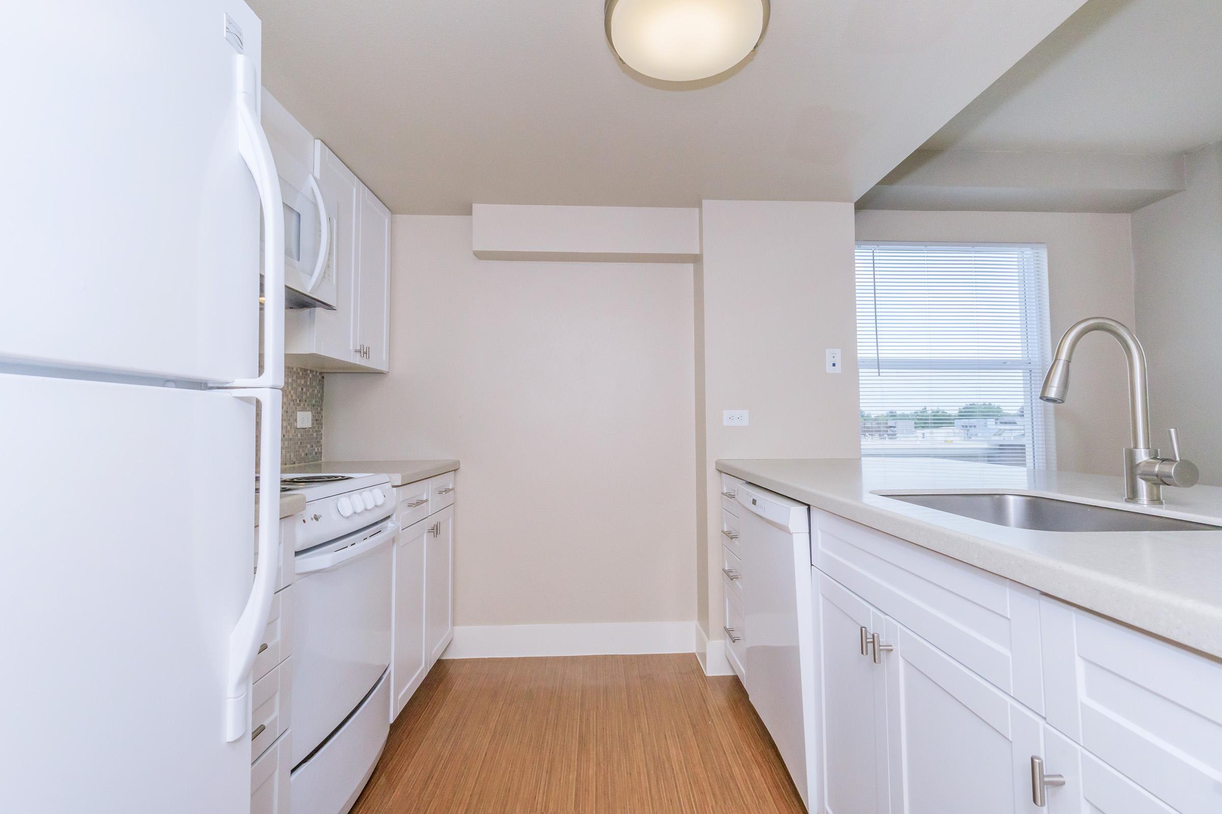 Bright, modern kitchen featuring white cabinetry, a stainless steel sink, and matching appliances, including a refrigerator and stove. The space has a light-colored wall and wood-style flooring, with a window allowing natural light to enter.