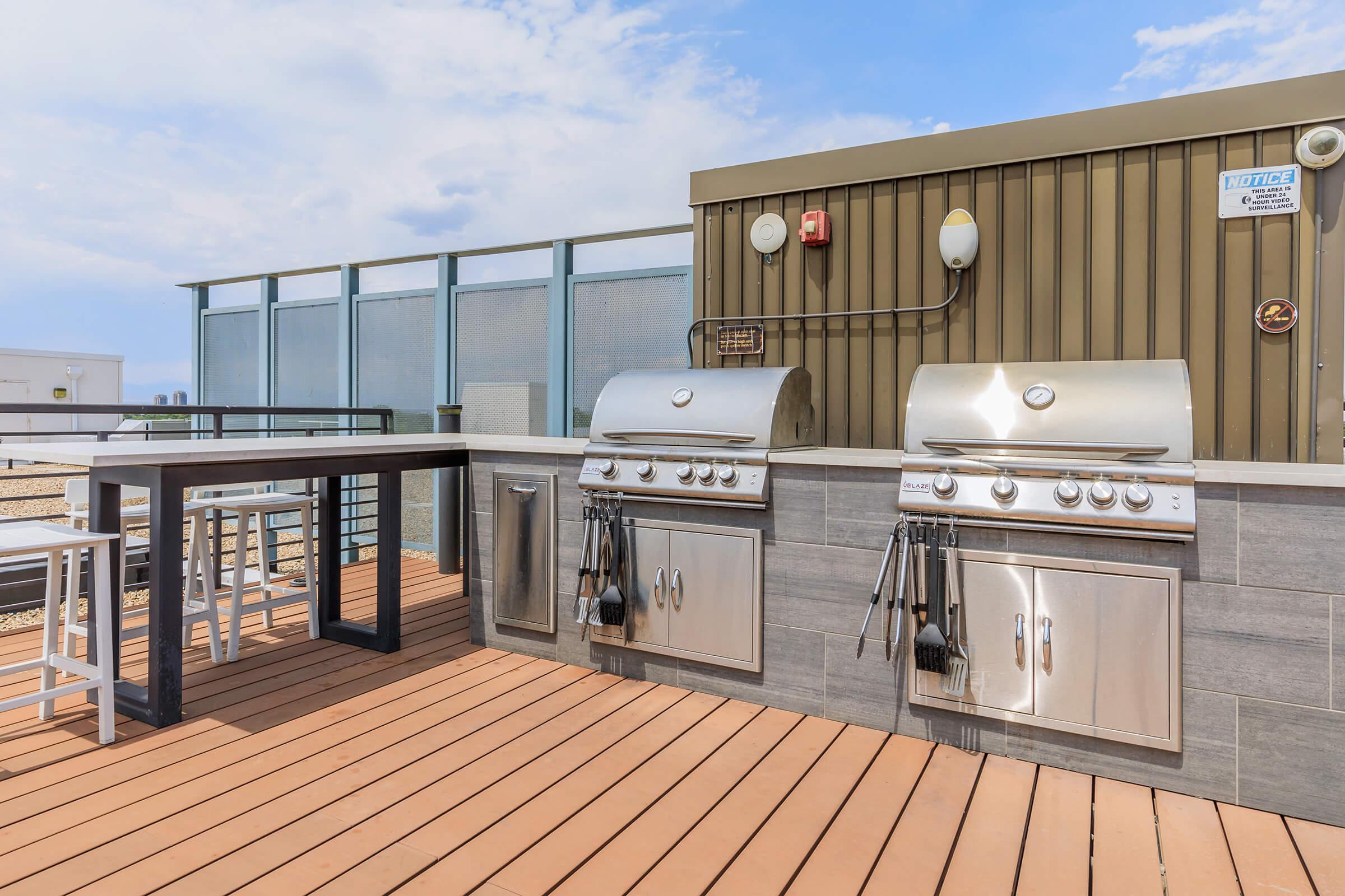 Outdoor kitchen area featuring two stainless steel gas grills on a wooden deck. There's a modern bar-height table with stools nearby. The backdrop includes a glass partition and a sky with light clouds, creating a spacious and inviting atmosphere for outdoor cooking and dining.