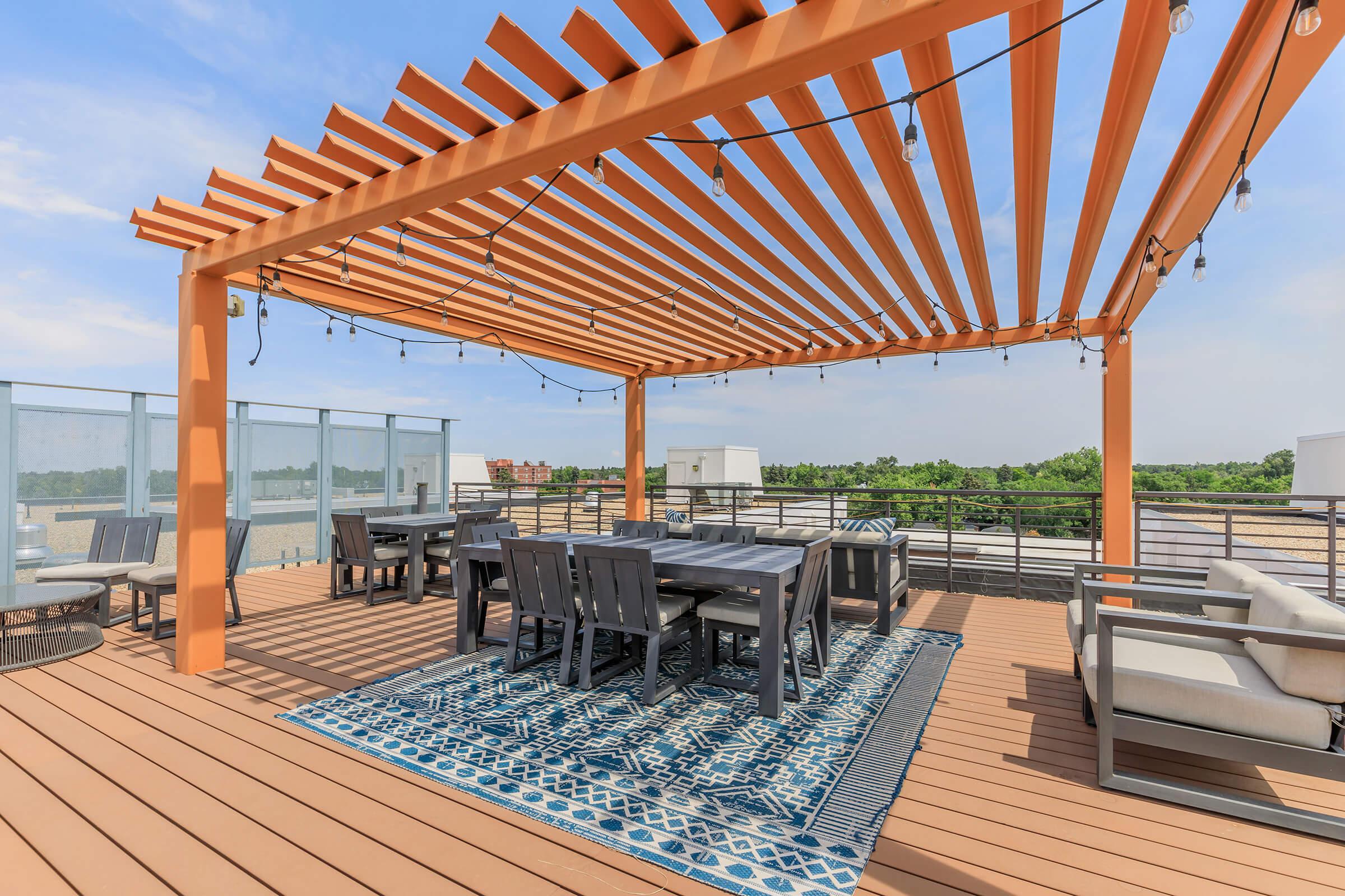A rooftop terrace featuring a wooden pergola with string lights, a large dining table surrounded by chairs, and comfortable seating arrangements. The space is adorned with a patterned area rug and offers a view of greenery in the distance, creating a cozy outdoor atmosphere.
