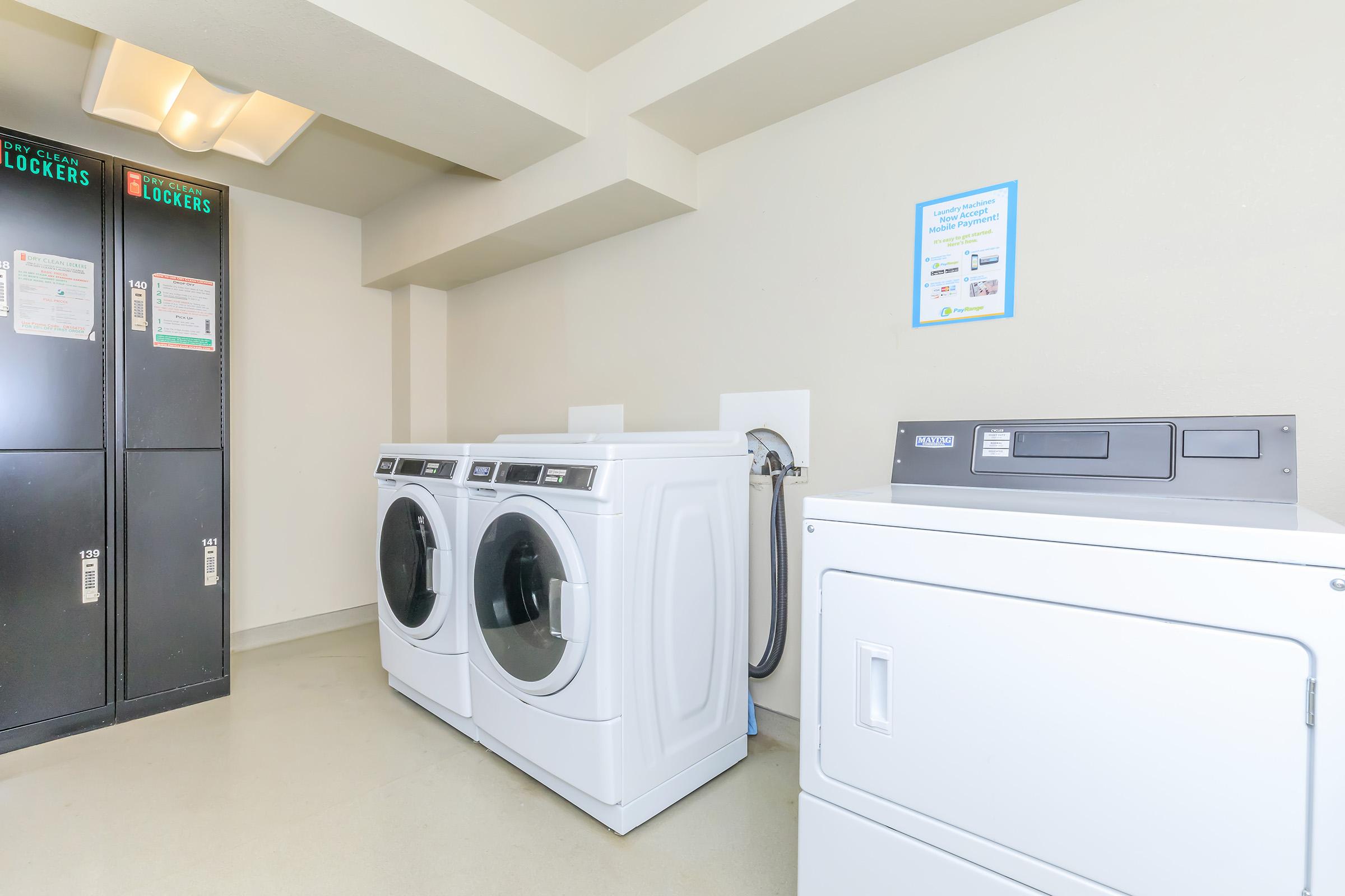 A laundry room featuring two large white washing machines and a white dryer. To the left, there is a gray locker unit with multiple compartments. The walls are painted a neutral color, and a small informational sign is posted nearby. The floor is clean and accommodates the laundry appliances.