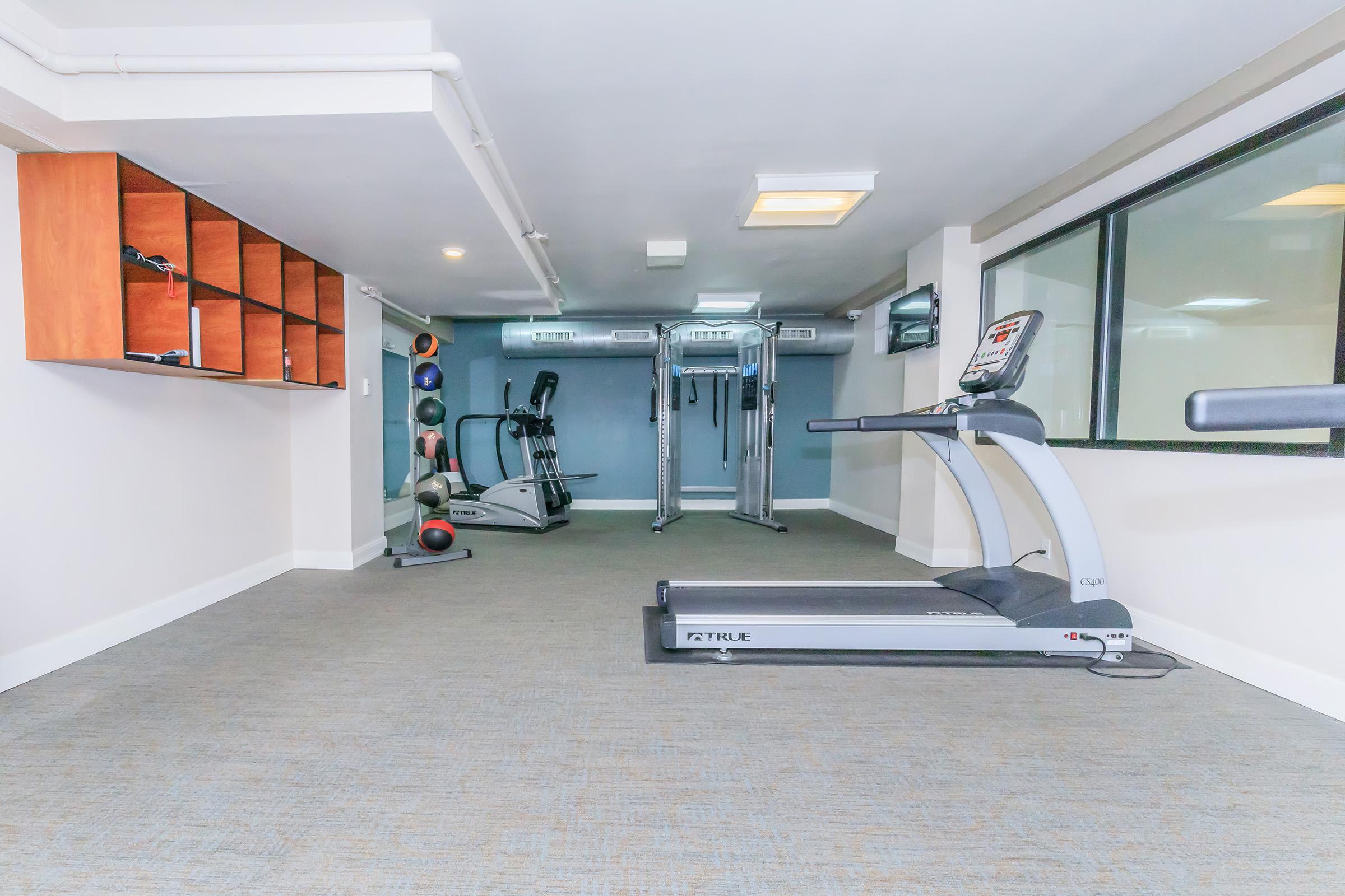 A well-equipped gym with a treadmill in the foreground, exercise machines in the background, and a variety of weights on a shelf. The space is bright and modern, featuring carpeted flooring and large windows that allow natural light to illuminate the area.