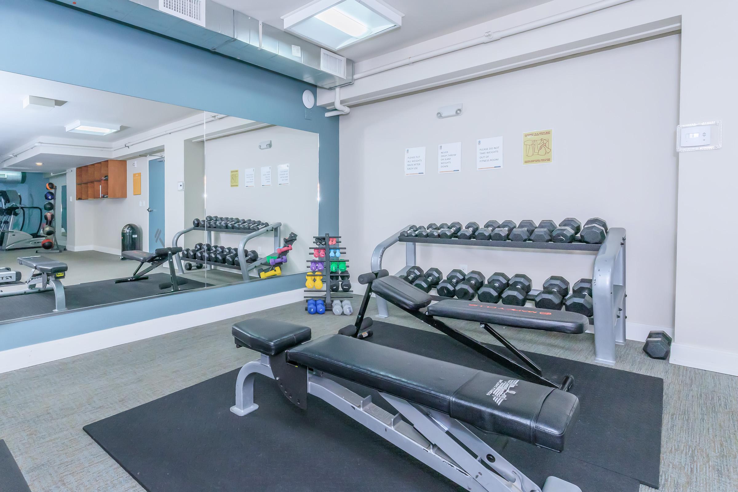 A fitness room featuring a bench, a rack of dumbbells in various colors, and a mirror reflecting the space. The floor is carpeted, and there are exercise machines in the background, contributing to a well-equipped workout area.