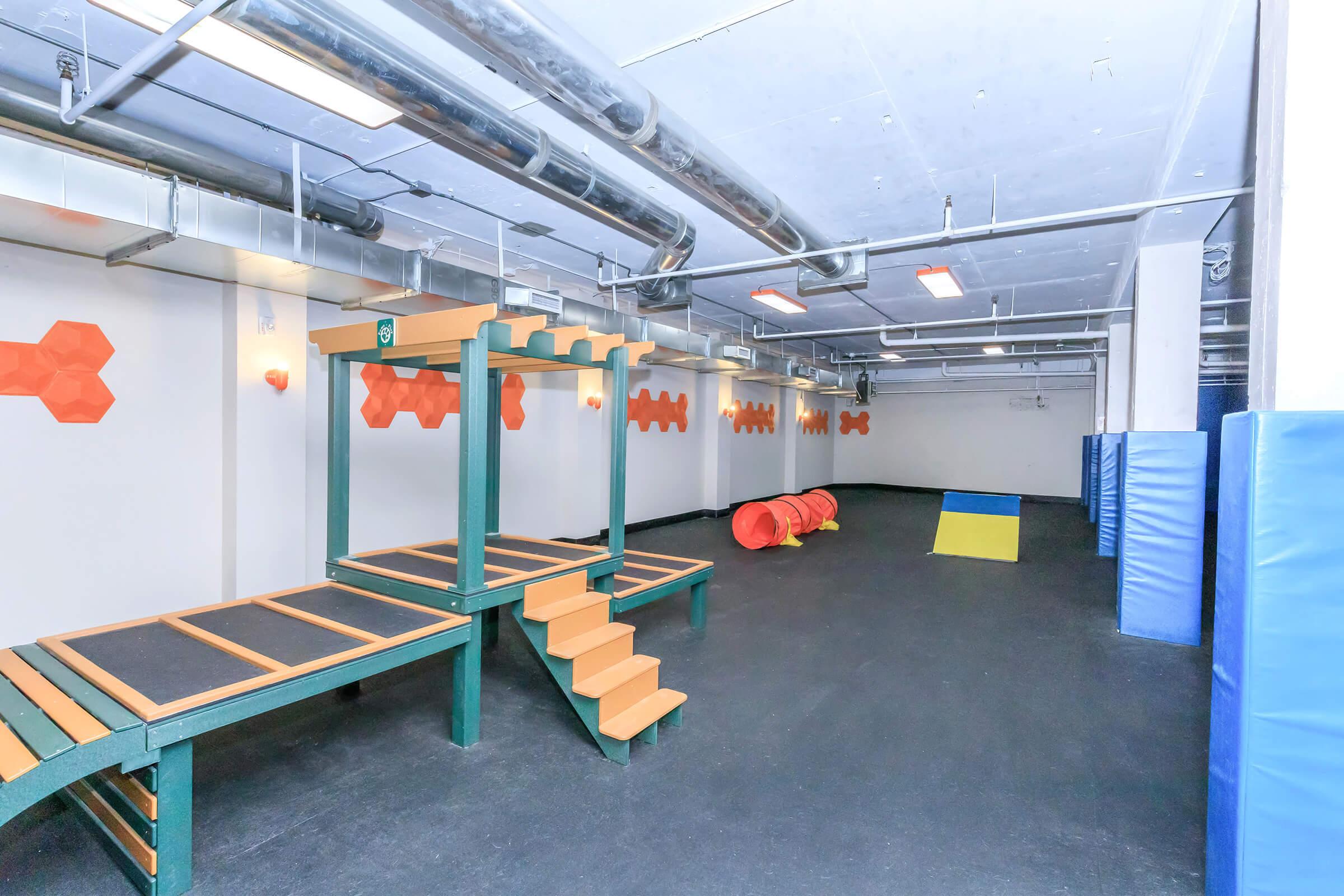 Indoor play area featuring multi-level climbing structures, a ramp, and colorful obstacles. Brightly lit space with padded flooring and orange wall decorations, suitable for children's activities and physical play.