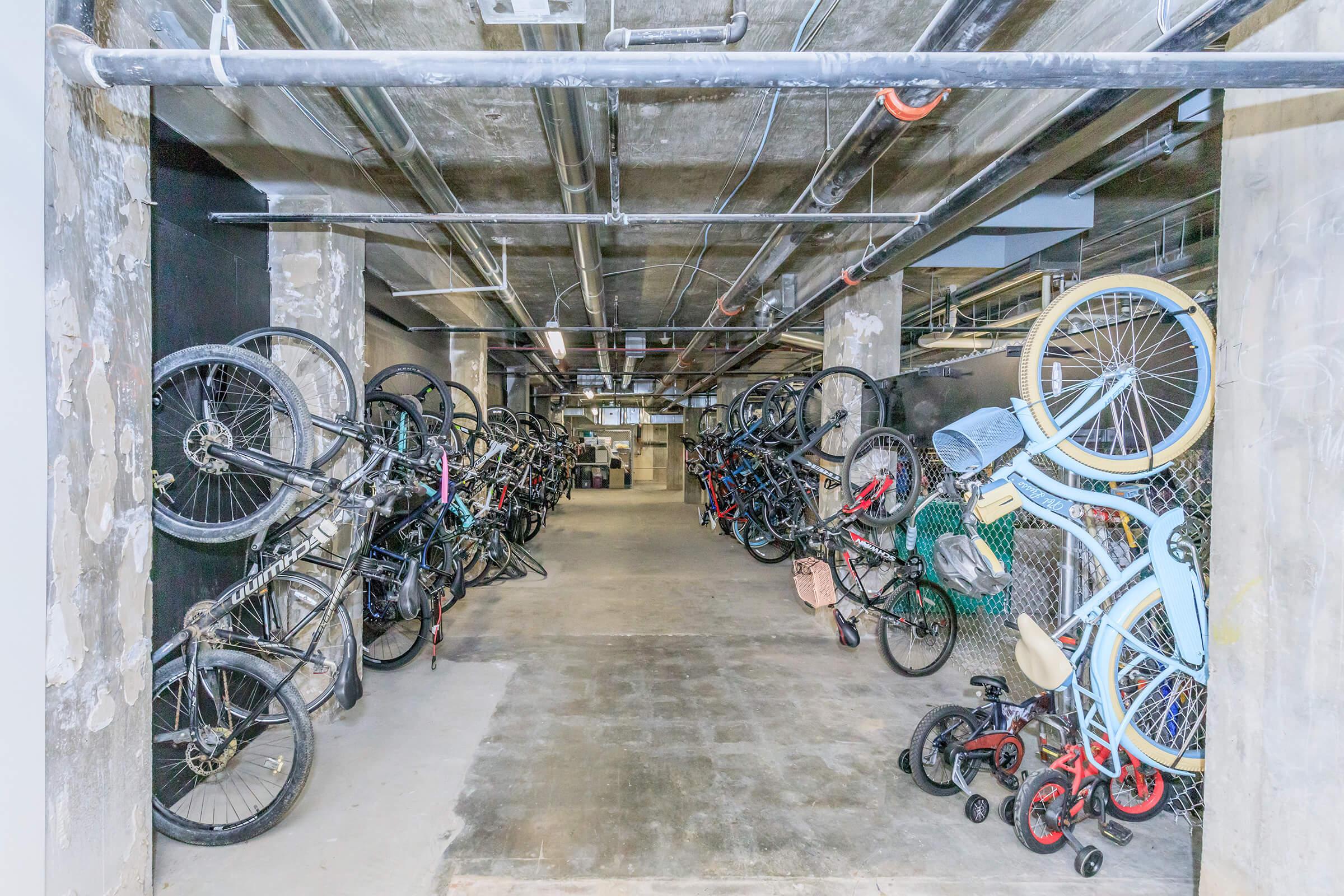 A dimly lit underground bike storage area featuring an array of bicycles leaning against walls and hanging from racks. The walls are marked with signs of wear, and overhead pipes run along the ceiling, creating an industrial atmosphere. Various styles and colors of bikes are visible, indicating a mix of uses.