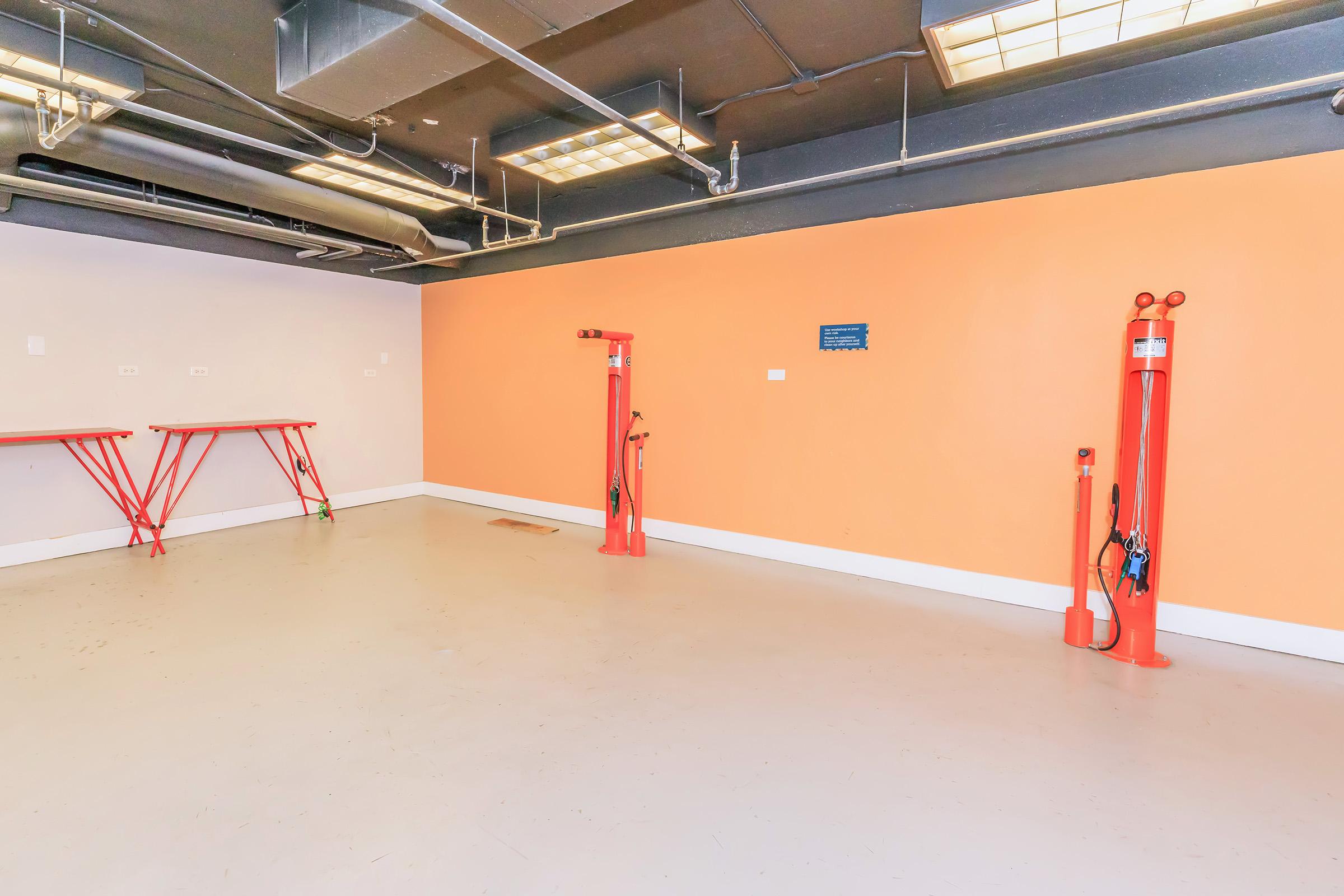 Empty room with an orange accent wall, featuring two red hydraulic lifts on the left and right. There are two red folding tables against the wall, and overhead lighting is provided by ceiling-mounted fixtures. The floor is light-colored and clean, creating an industrial space.