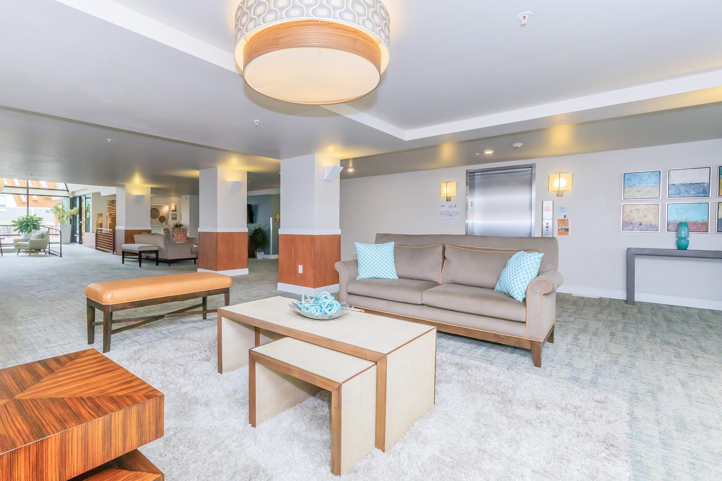 A modern lobby area featuring a comfortable brown sofa with light blue cushions, a wooden coffee table, and a bench. The space has neutral-colored carpeting and decorative wall art, along with an elevator in the background. Soft lighting creates a welcoming atmosphere with plants adding greenery.