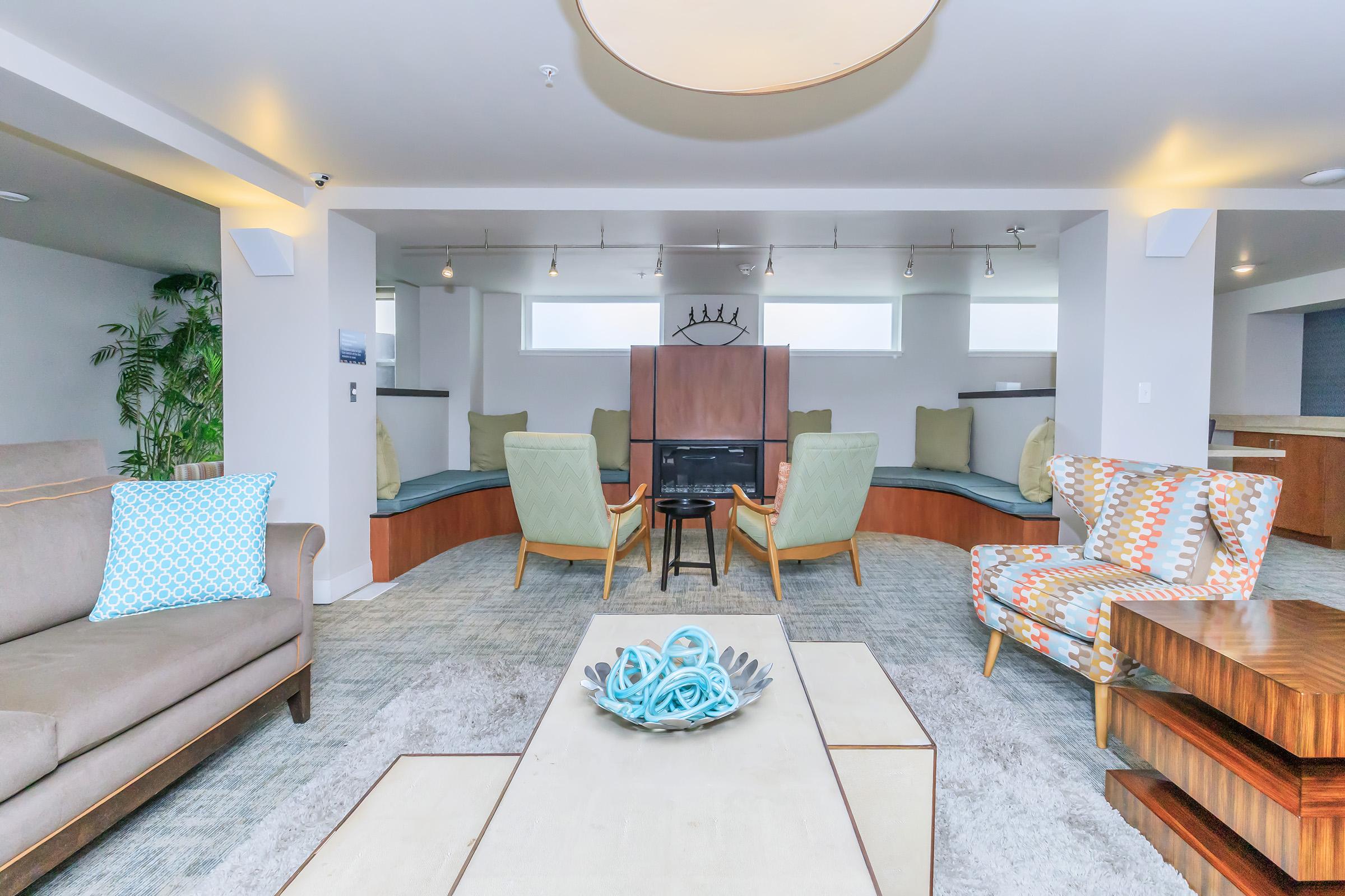 A modern living area featuring comfortable seating with a light color palette. There are two patterned chairs, a sofa, and a low coffee table with decorative items. The space includes built-in seating with cushions, plants, and large windows, creating an inviting and cozy atmosphere.