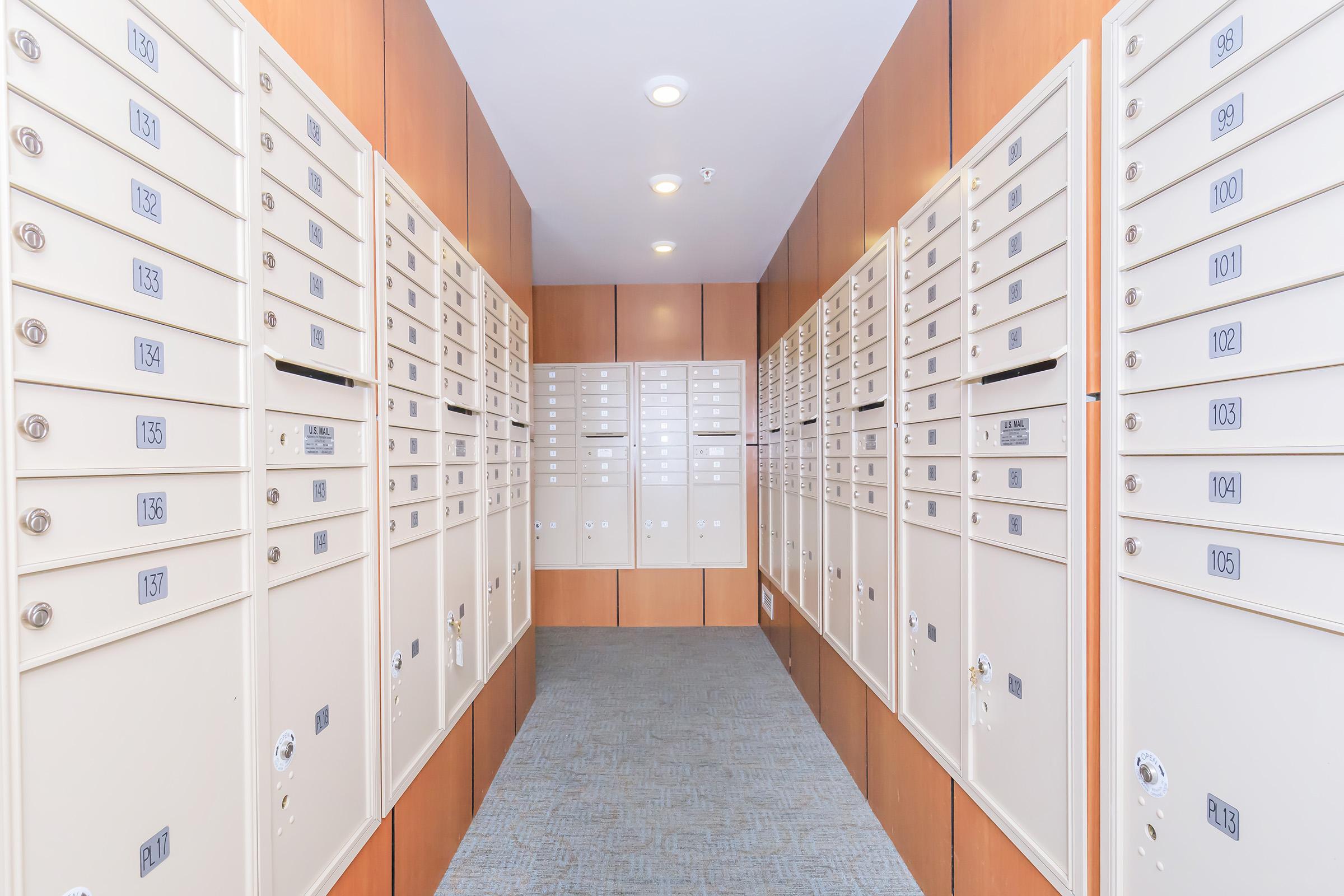 A corridor lined with multiple rows of mailboxes. The walls are wooden, and the mailboxes are light-colored with numbered compartments. The floor is covered with gray carpet, and overhead lights illuminate the space, creating a clean and organized environment for mail collection.