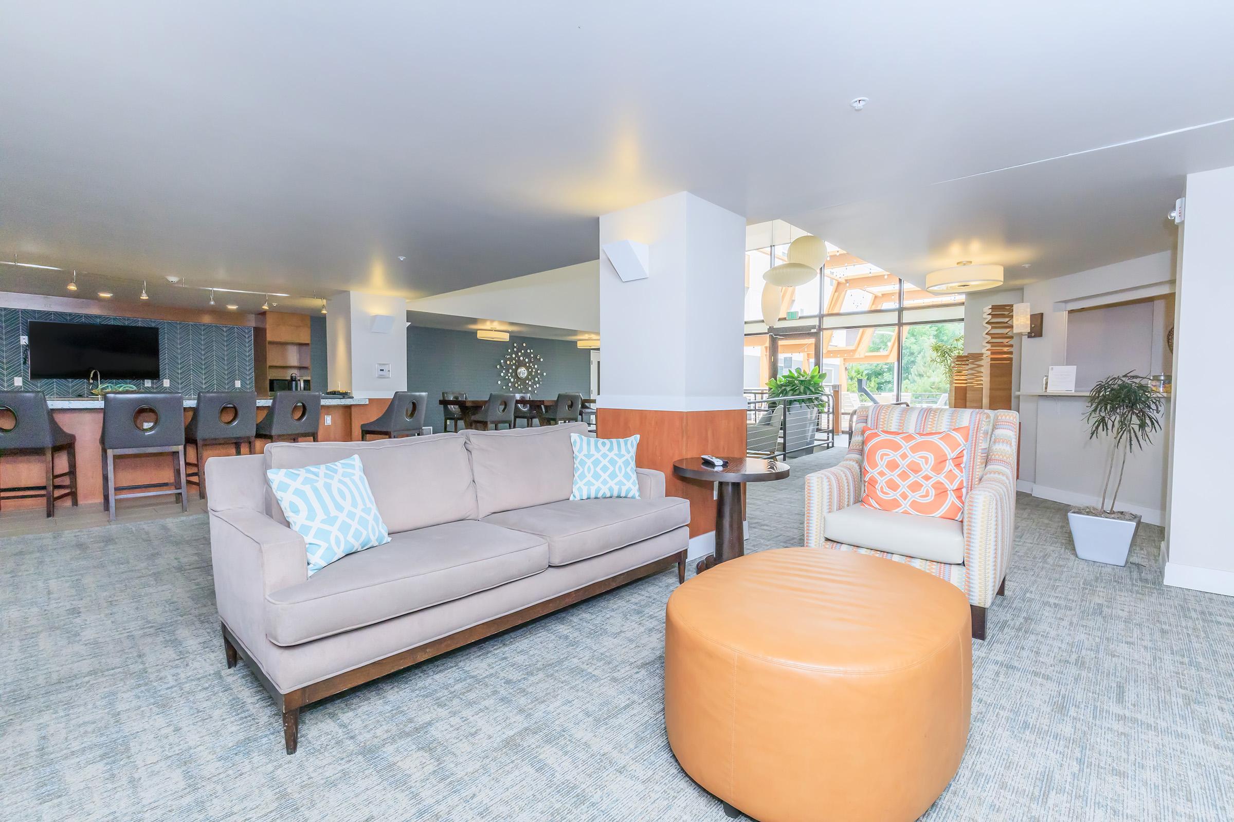 A modern lounge area featuring a comfortable gray sofa with decorative pillows, an orange upholstered ottoman, and a patterned armchair. The space includes a bar with stools in the background and large windows that let in natural light, creating a welcoming atmosphere.