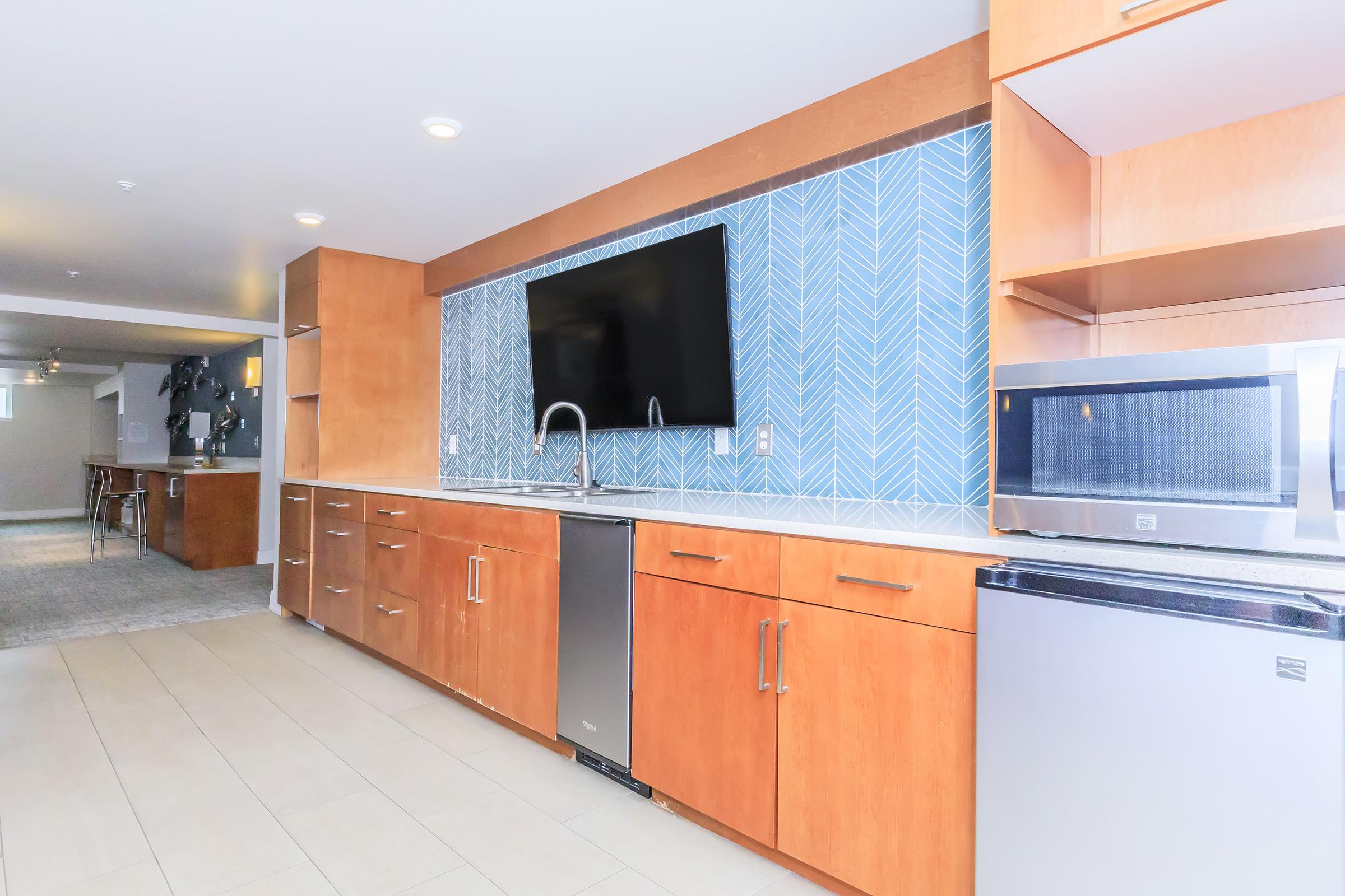 Modern kitchen interior featuring wooden cabinetry, a large flat-screen TV mounted on a blue herringbone tile wall, a stainless steel sink, and appliances including a microwave and a dishwasher. Bright lighting enhances the clean, contemporary design.