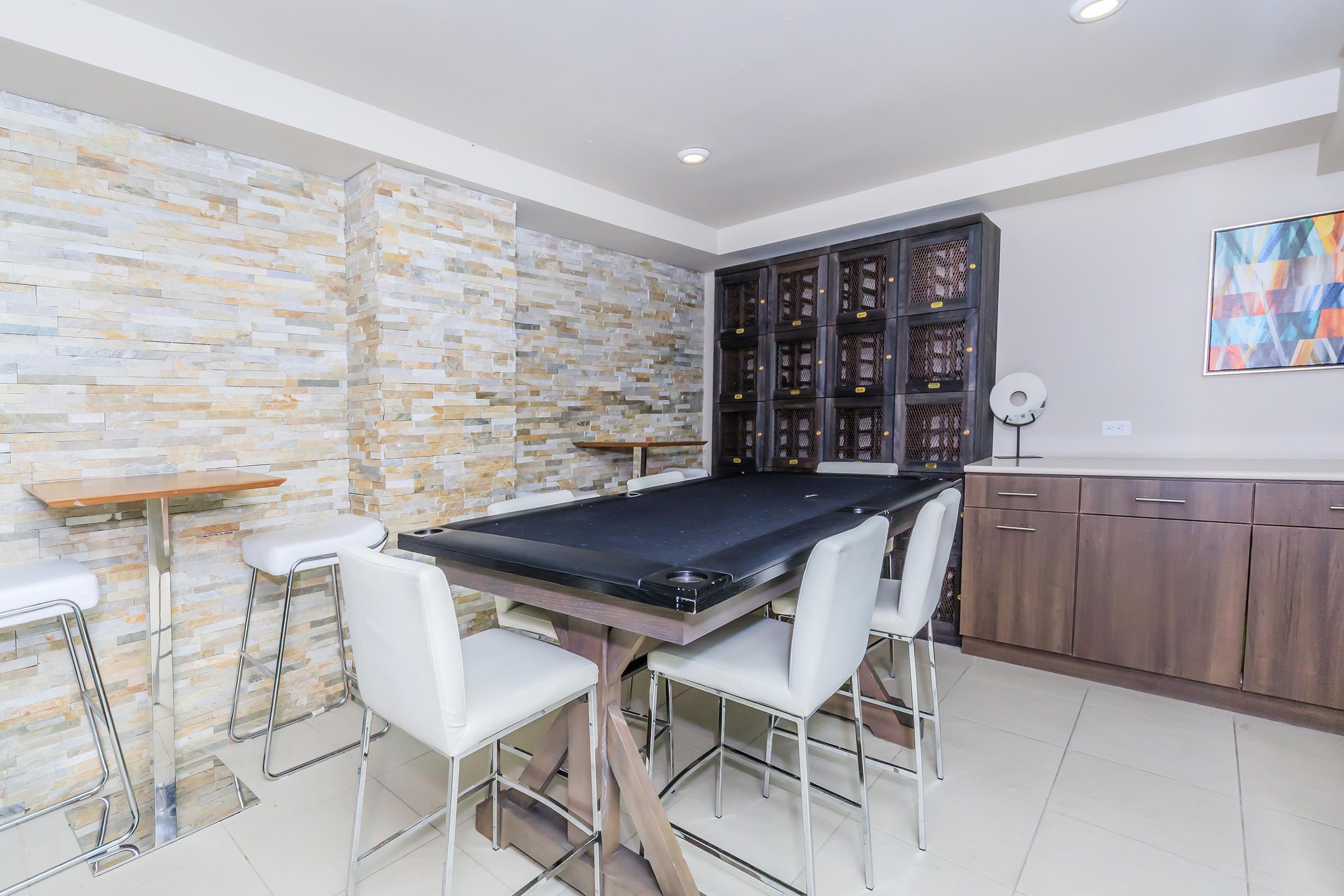 A modern dining area featuring a sleek wooden table with a black tabletop surrounded by white high-back chairs. The room is accented with a textured stone wall and a stylish wine cabinet, complemented by minimalist bar shelves and an abstract artwork on the wall. Bright lighting enhances the contemporary design.