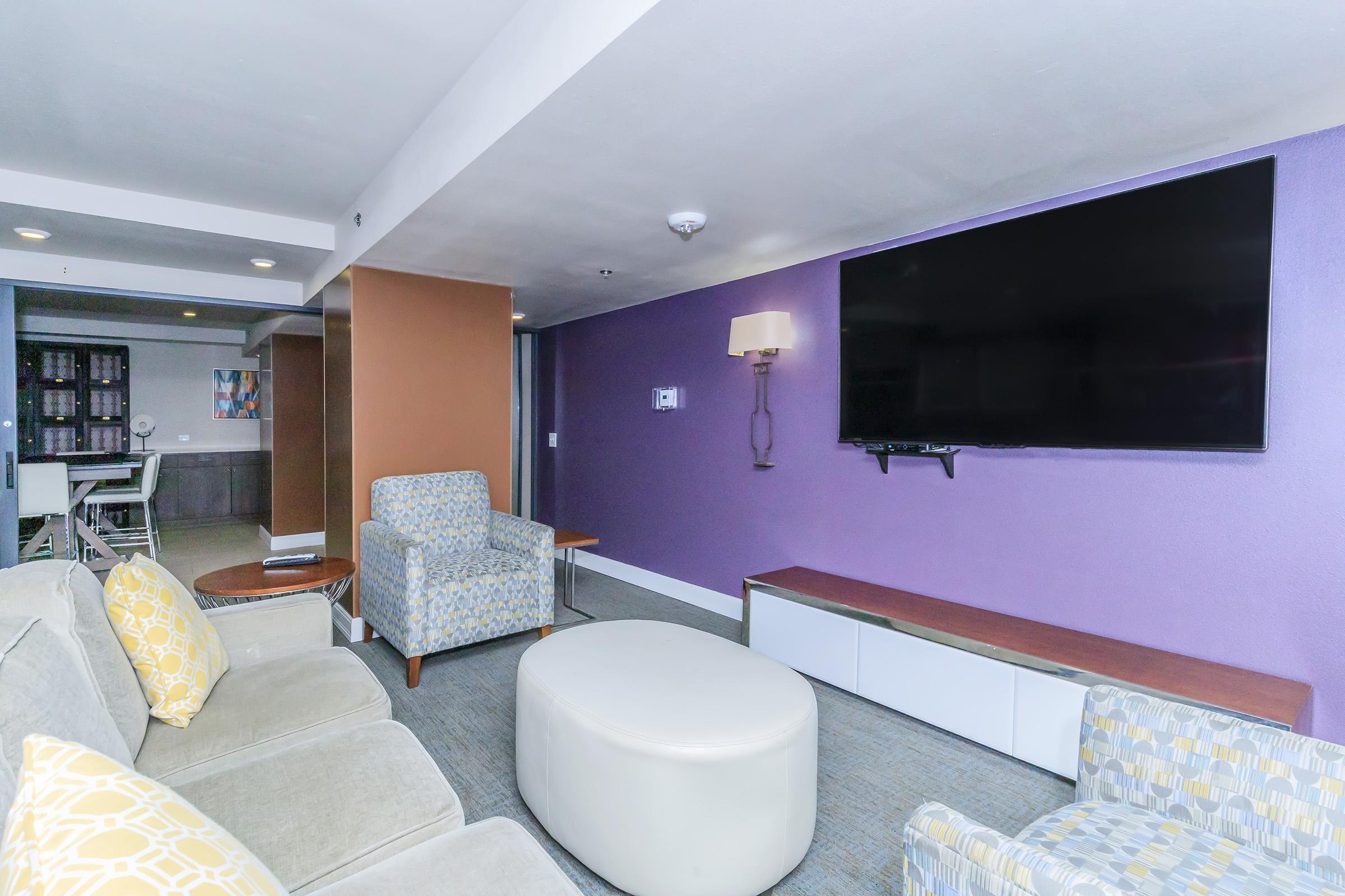 Modern lounge area featuring a large flat-screen TV mounted on a purple wall, comfortable light-colored seating with decorative pillows, a round white coffee table, and a cozy atmosphere. A lamp provides additional lighting, and there is a glimpse of a kitchen or dining area in the background.
