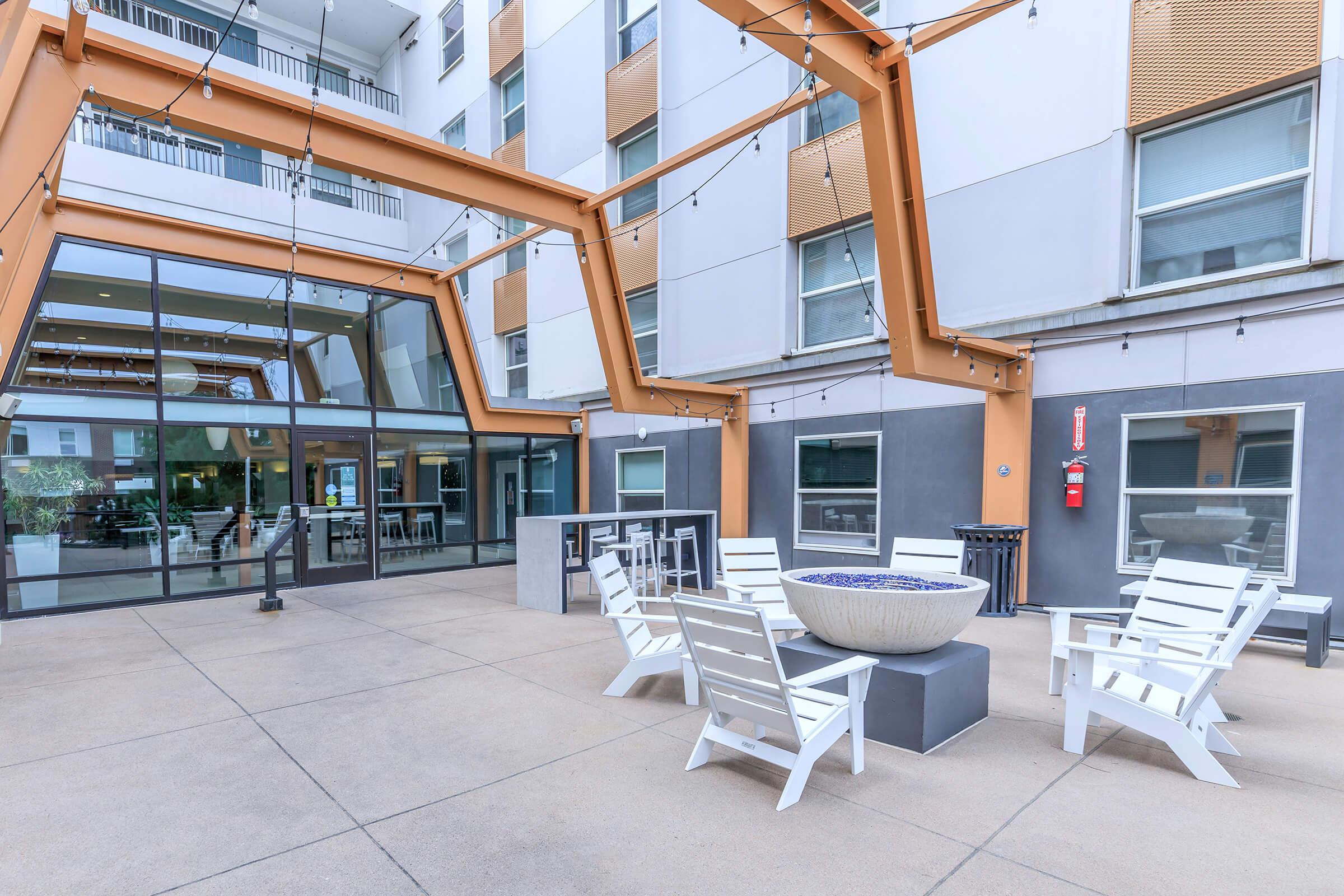 Outdoor lounge area featuring white chairs and a circular fire pit, with modern architecture in the background. Large glass windows provide a view into a communal area. The space is designed for relaxation and social gatherings, highlighted by geometric beams and ambient lighting.