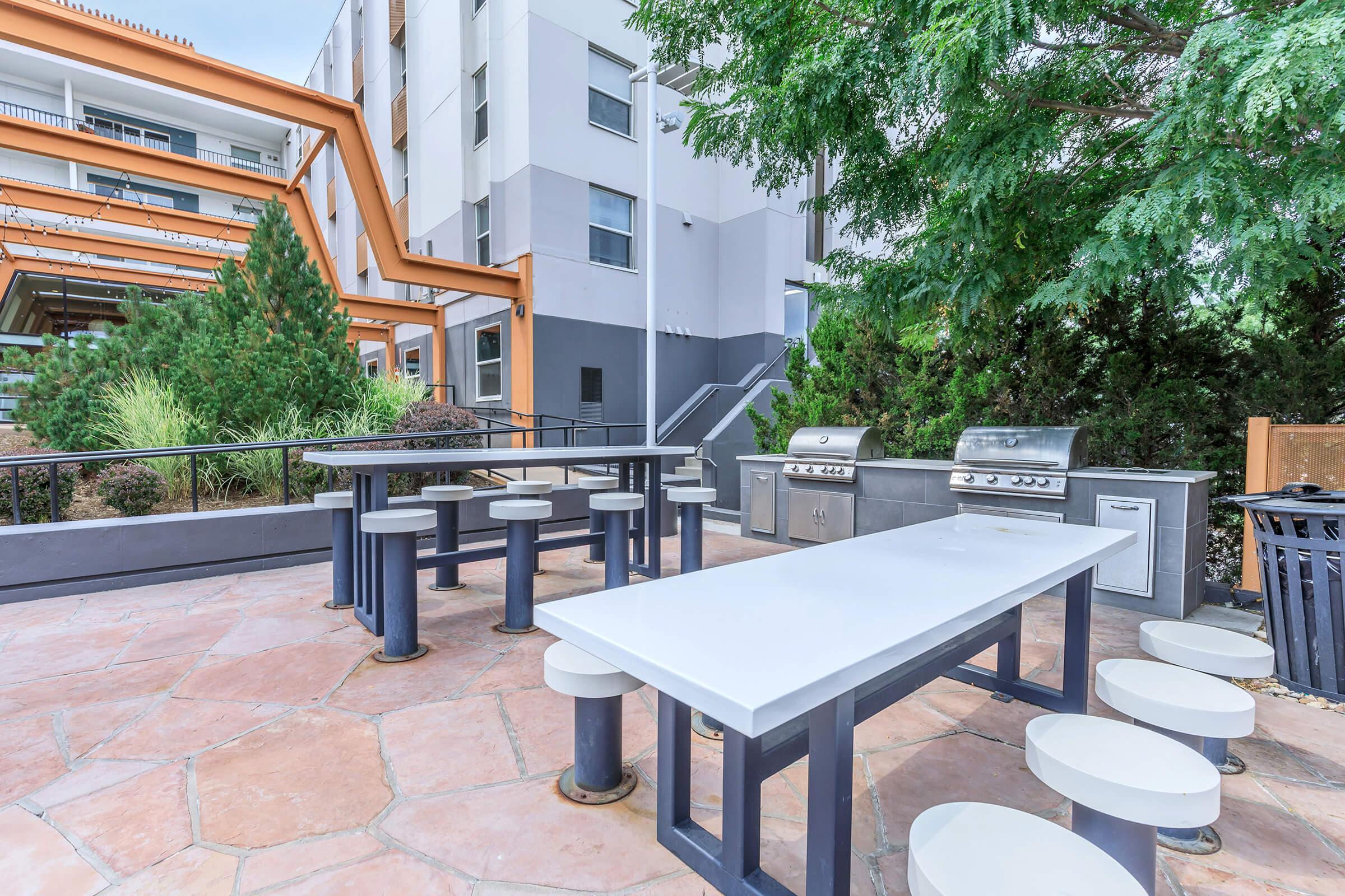 Outdoor grilling area featuring two barbecue grills, several tables, and surrounding greenery. The area is well-maintained, with a modern design and ample seating, ideal for gatherings and cookouts. Buildings are in the background, complemented by trees for shade.