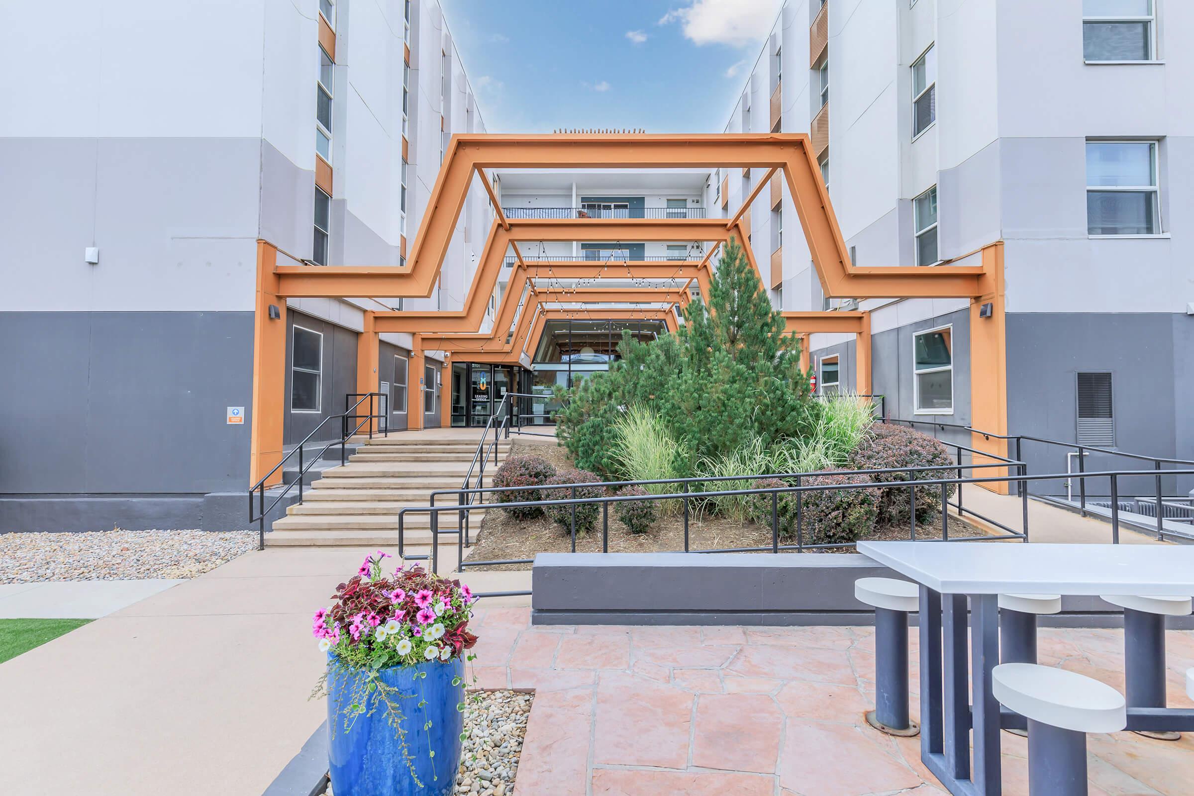 Modern apartment building entrance with a distinctive architectural overhang, featuring lush greenery and landscaped areas. A set of stairs leads up to the main entrance, while a seating area with a table is visible in the foreground, surrounded by colorful flower pots. Clear blue sky in the background.