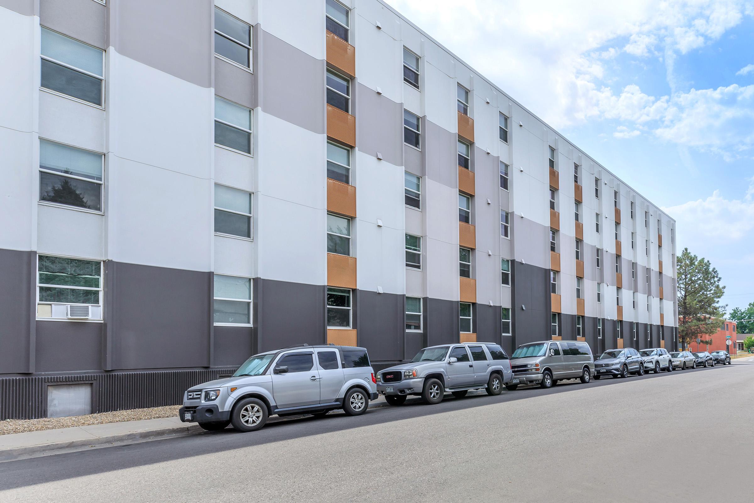 A modern multi-story building with a mix of beige, gray, and orange panels. Several parked vehicles line the street in front of the building, with blue skies and clouds visible above. The scene reflects a contemporary urban setting.