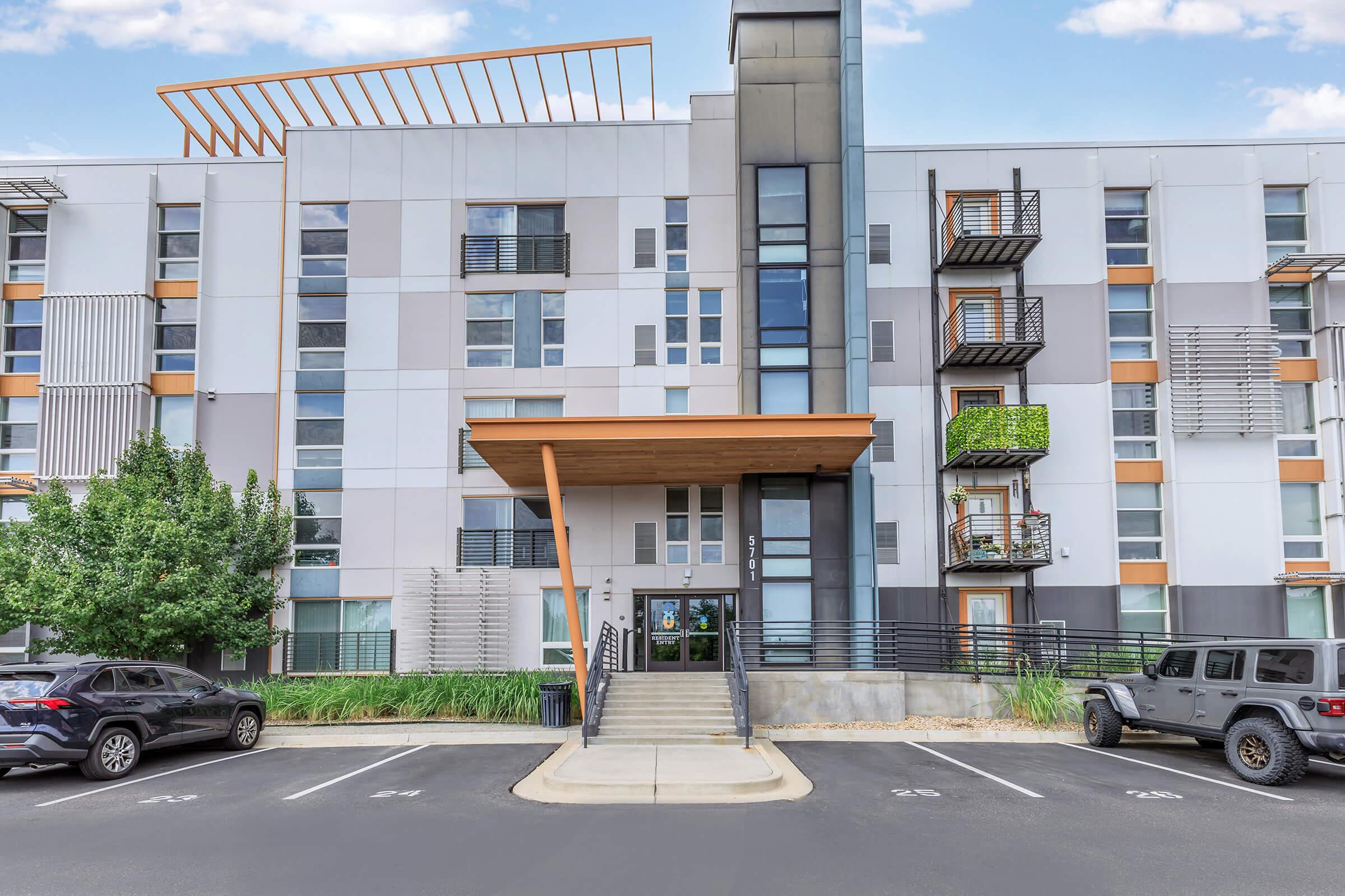 Modern multi-story residential building with a mix of gray and white exterior. The entrance features a wooden overhang and glass doors, flanked by balconies. Two parked cars are visible in front, with greenery around the entrance area. The overall design is contemporary and stylish.