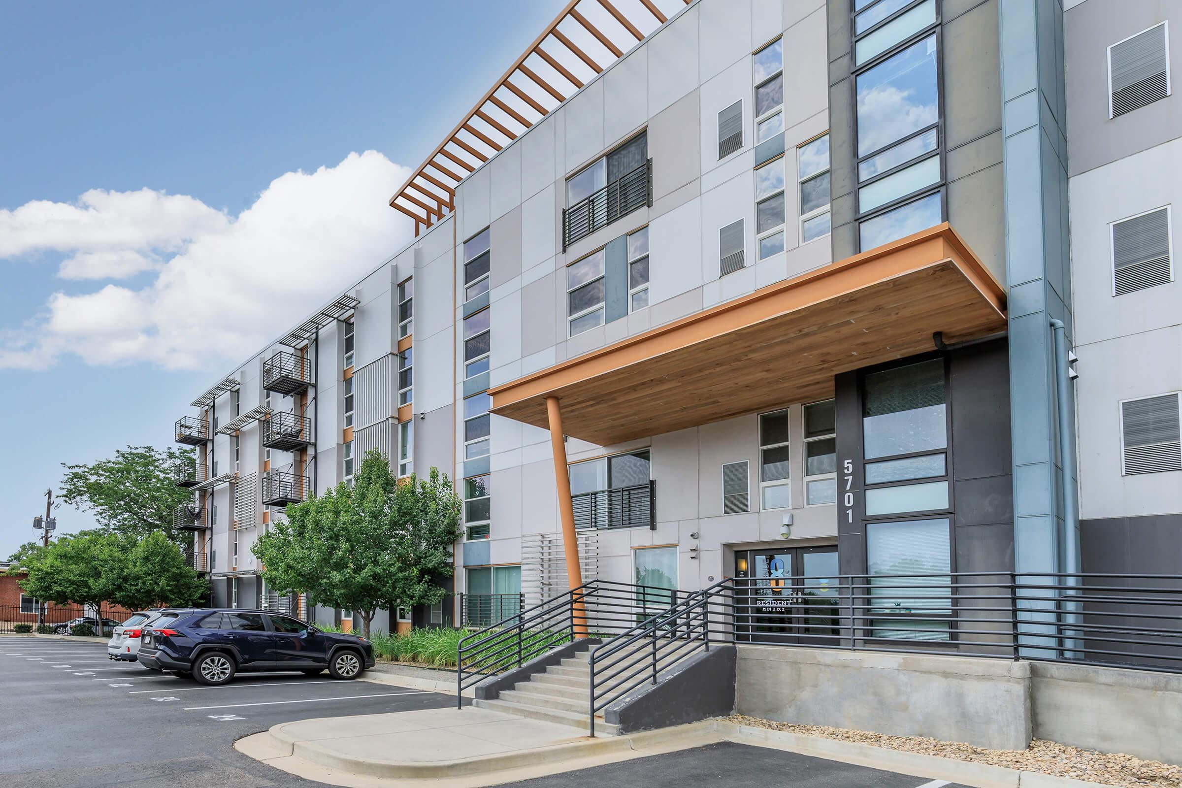 Modern apartment building with a sleek design featuring a wood-paneled entrance, large windows, and lush green landscaping. The exterior has a combination of light and dark materials, with several parked cars in front and blue sky with clouds above.