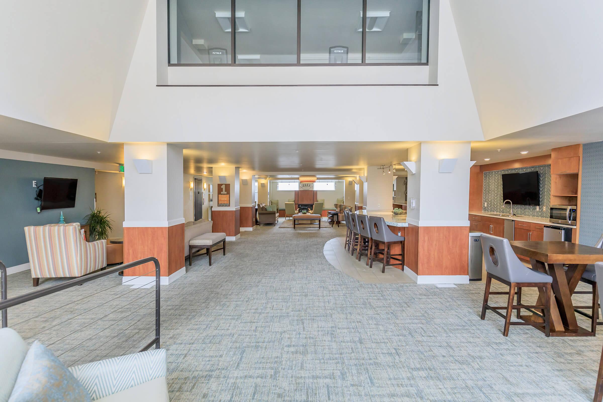 A bright, open common area featuring modern furnishings including seating arrangements, a dining table, and kitchen space. Large windows provide natural light, and the decor is neutral with soft colors, creating a welcoming atmosphere for social gatherings or relaxation.